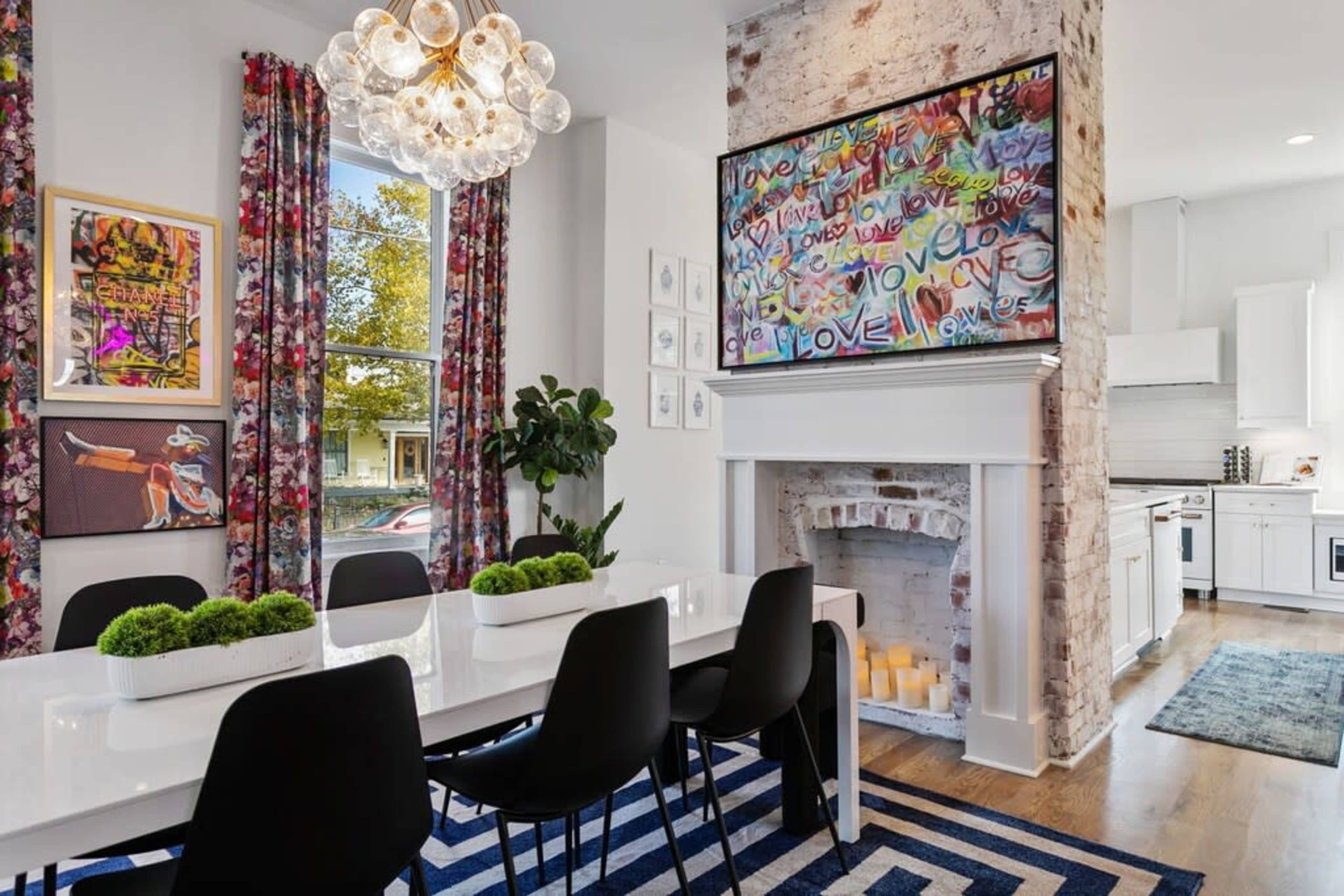 A modern dining area features a white table surrounded by black chairs, colorful curtains, and a large TV mounted above a brick fireplace.