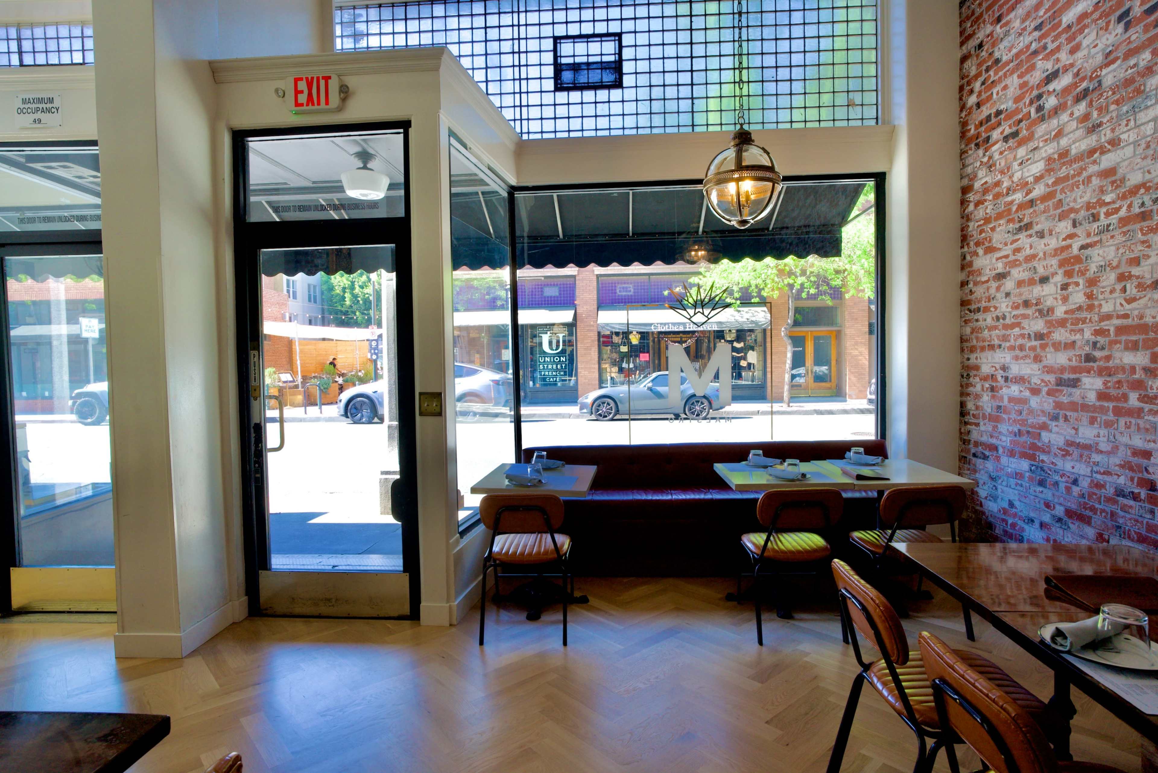 The image shows the interior of a restaurant featuring a large glass entrance, a brick wall, and tables set for dining.