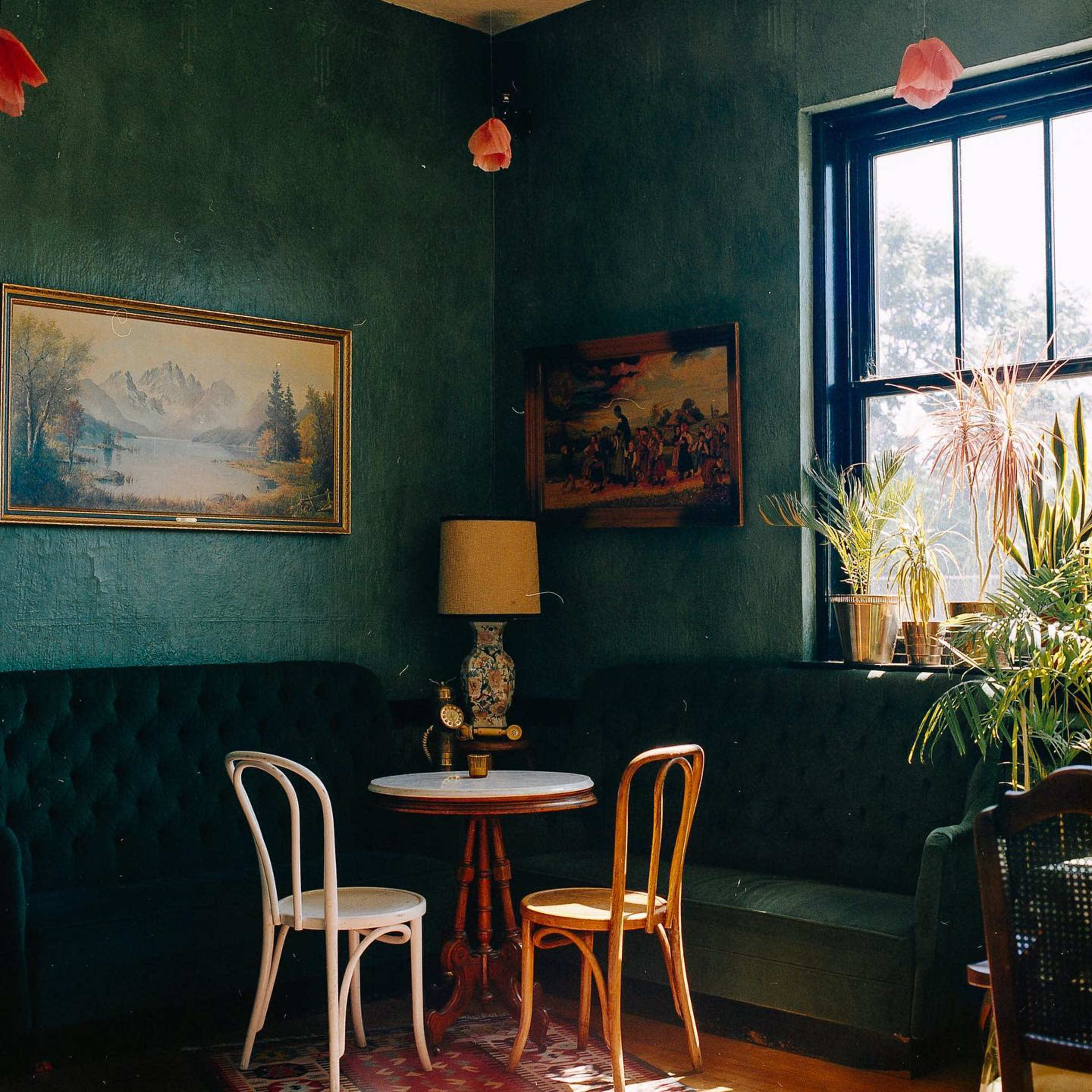 A cozy corner of a room featuring green walls, a round table with two chairs, a lamp, and framed paintings.
