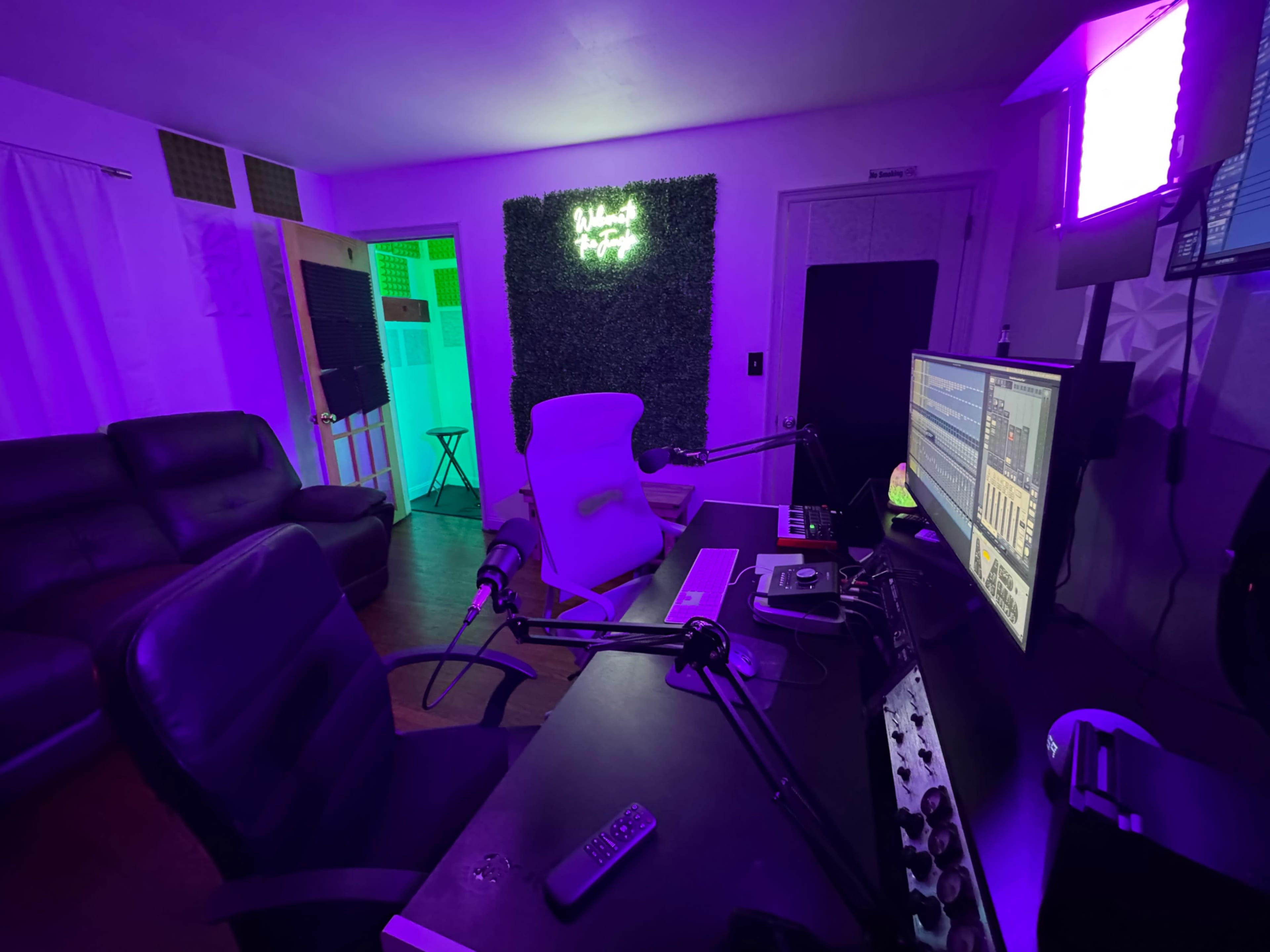 The image shows a dimly lit recording studio with purple lighting, a desk equipped with monitors and microphones, and a green neon sign on the wall.