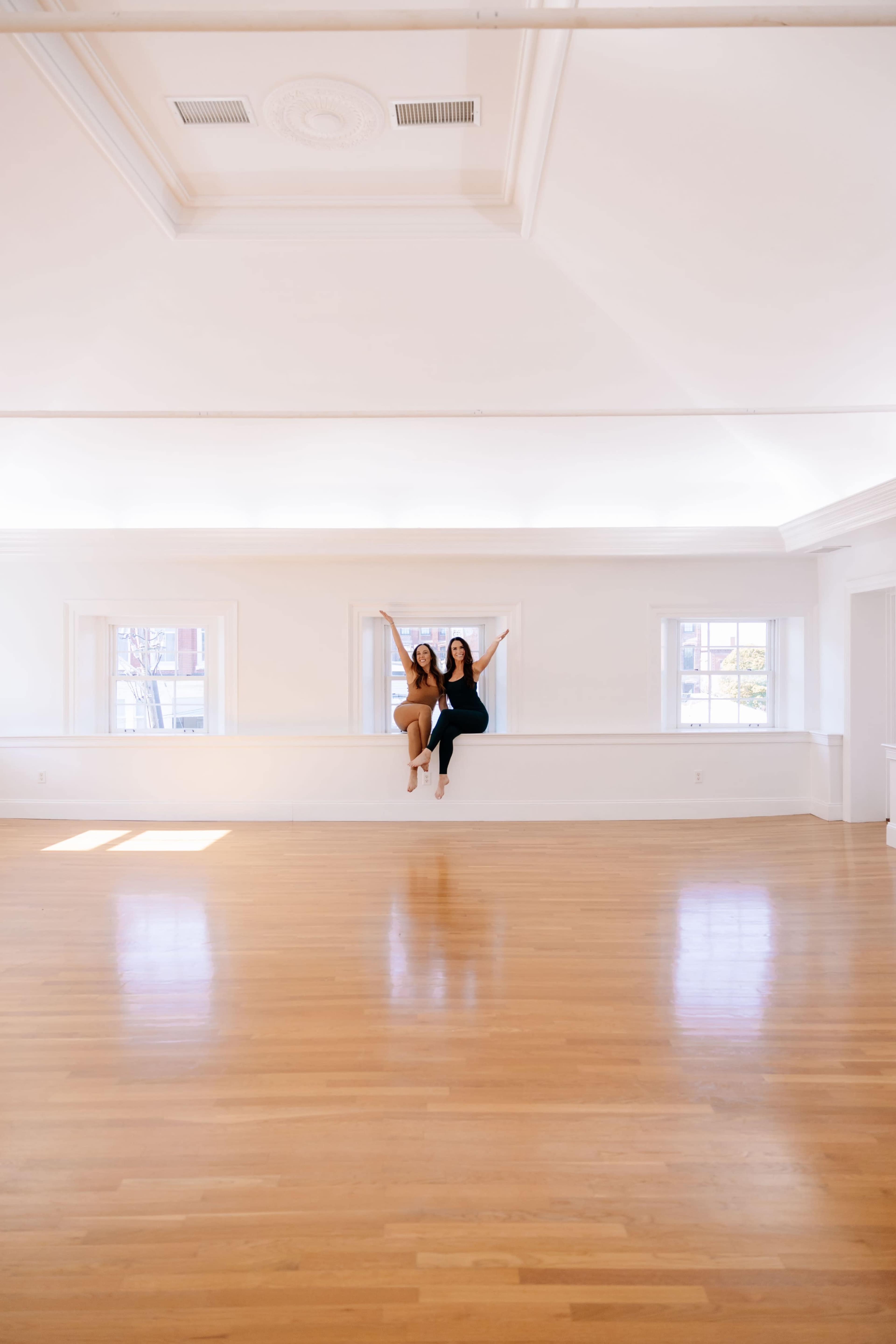 Two women sit on a ledge in a spacious, brightly lit room with wooden floors and large windows.