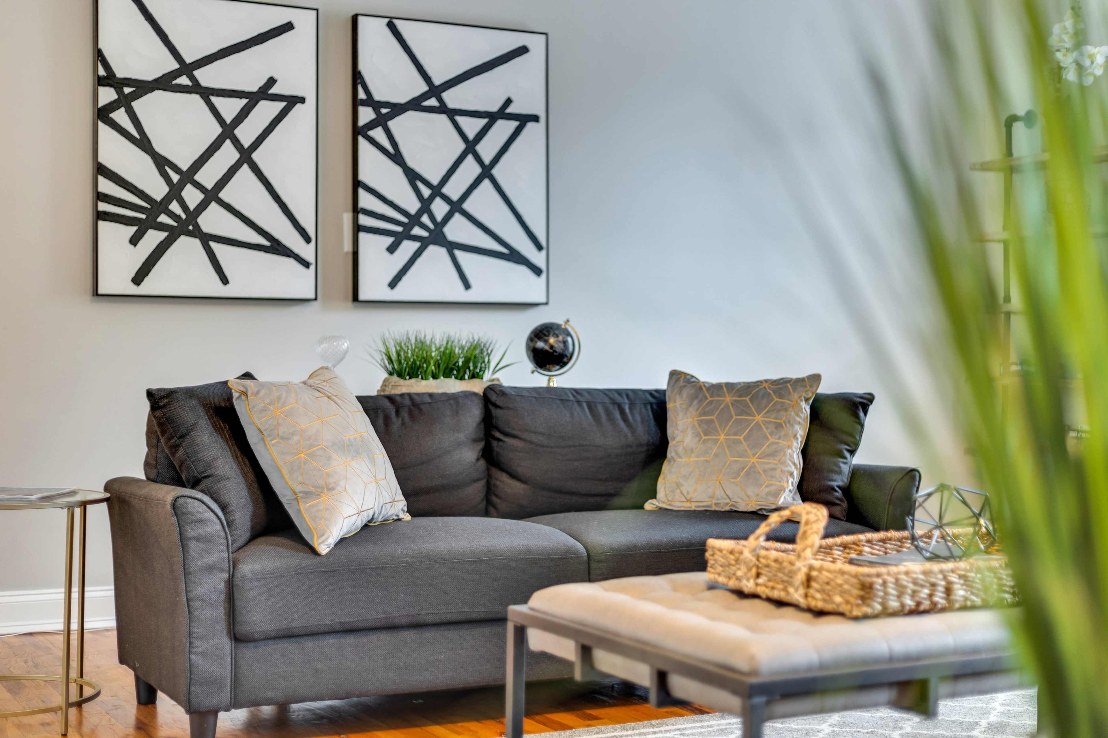 A gray sofa with decorative pillows sits in a living room adorned with abstract art on the walls and a coffee table with a woven basket.