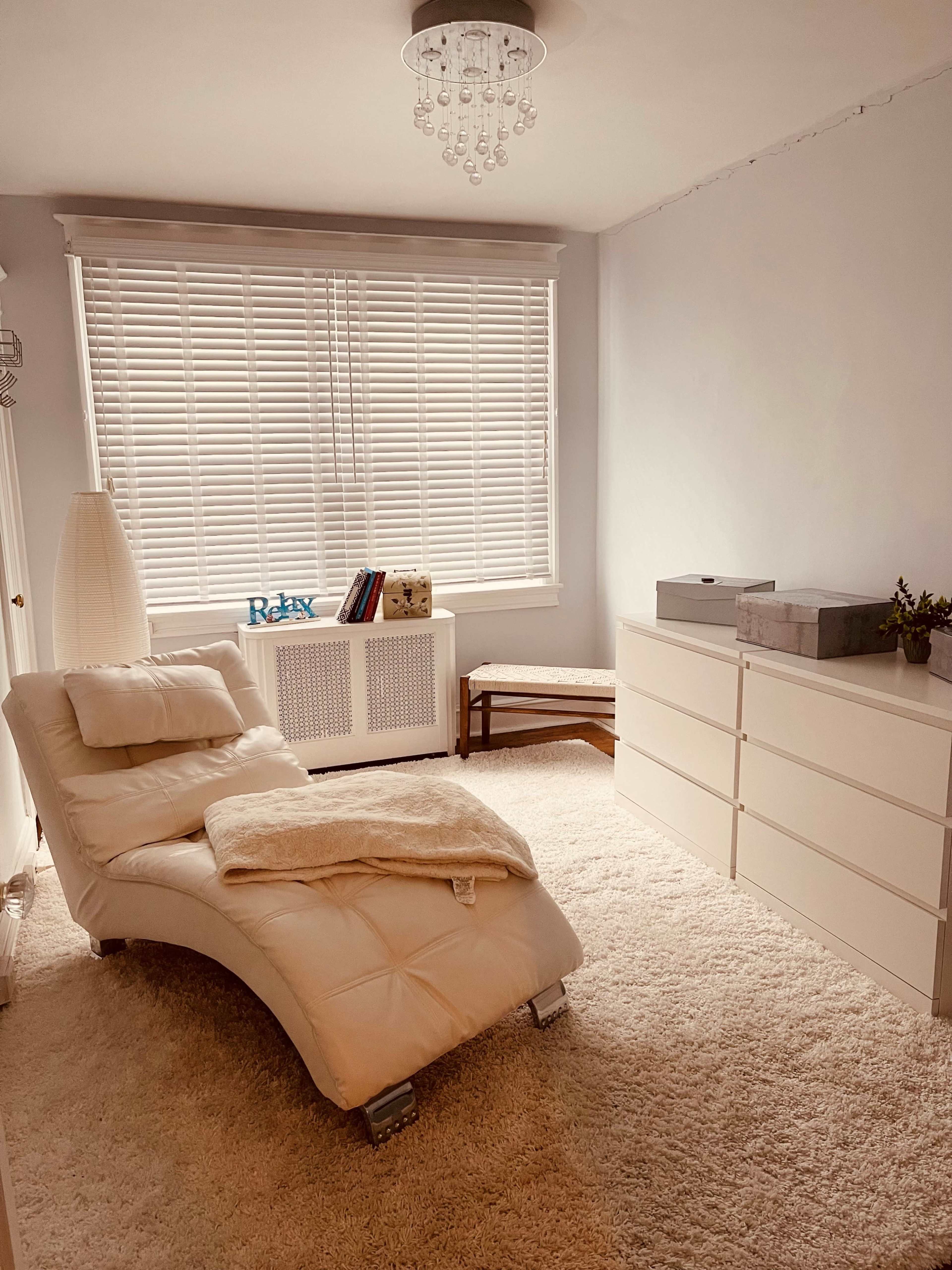 A light-colored room with a chaise lounge, a small bench, a dresser, and a window with white blinds.