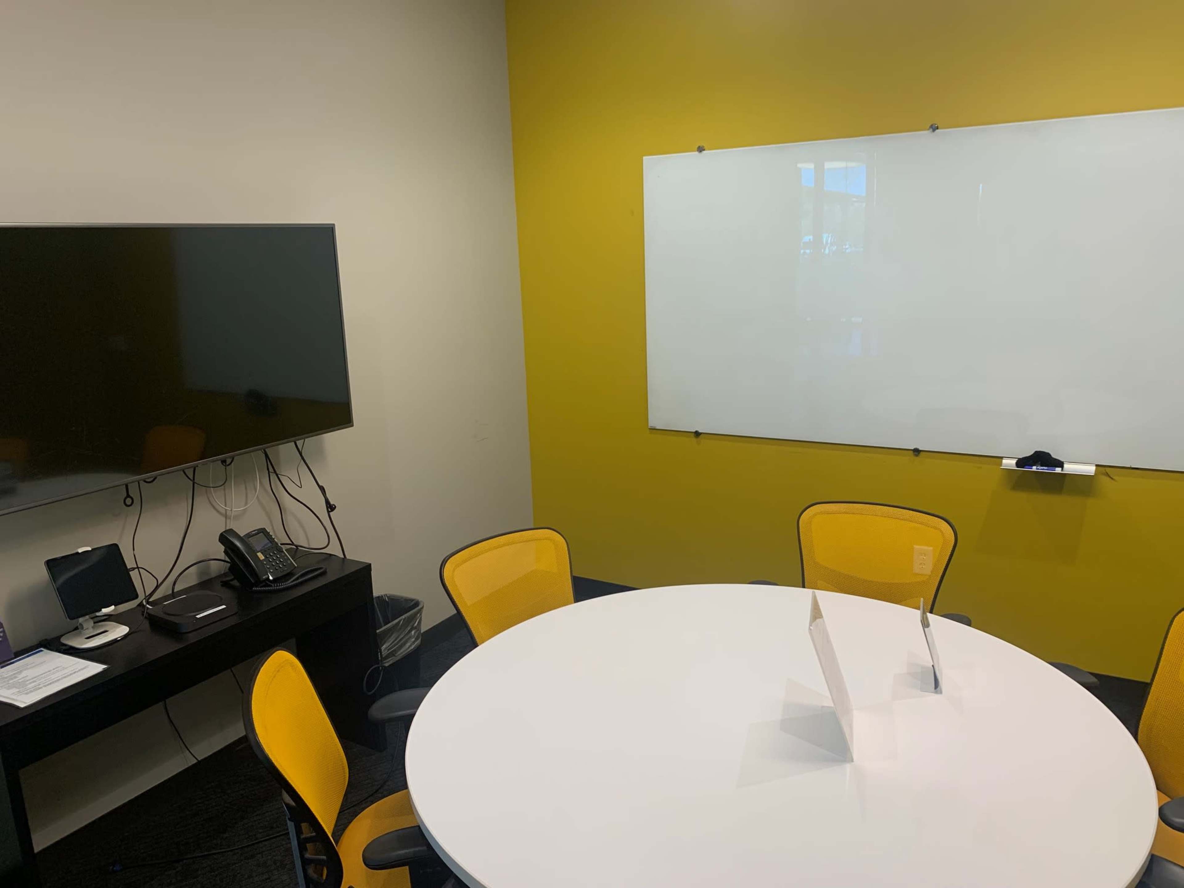 A conference room features a round white table, yellow chairs, a television mounted on the wall, and a whiteboard.