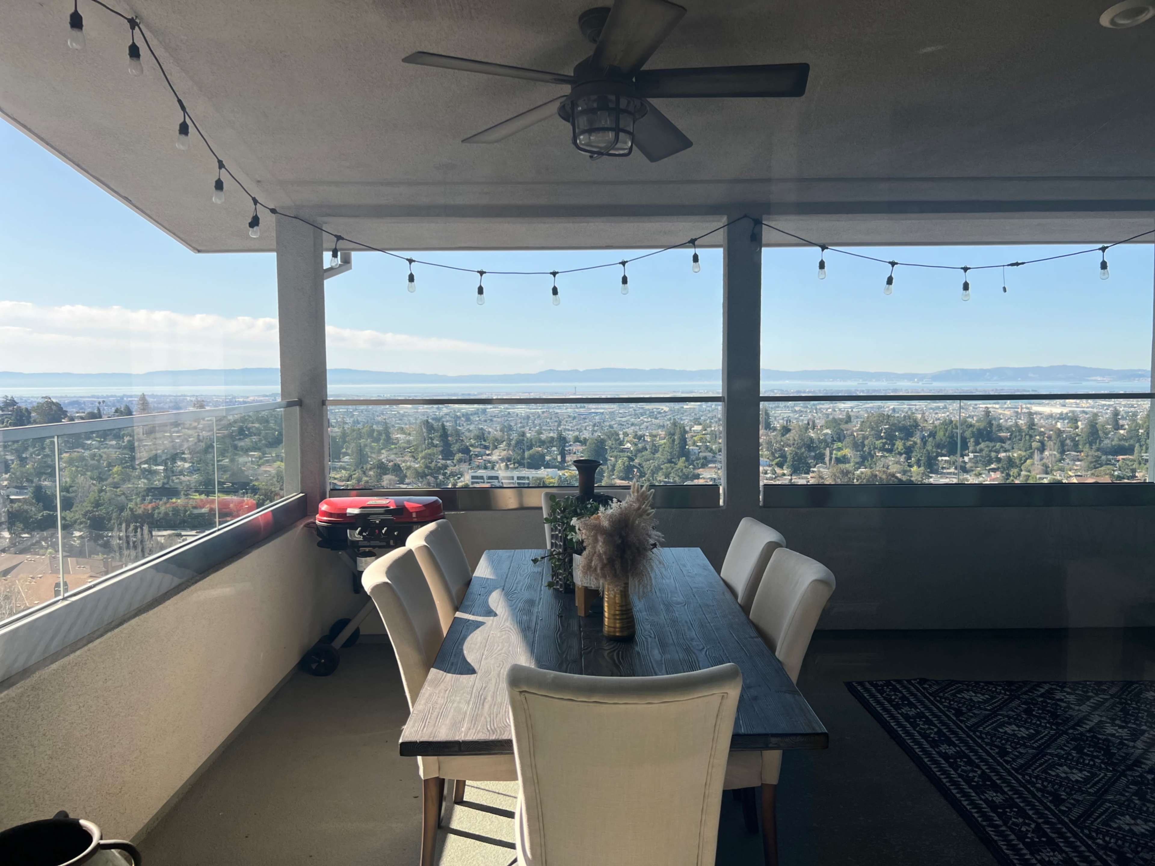 A dining table with six chairs is set on a balcony overlooking a cityscape and distant mountains, with string lights hanging above.
