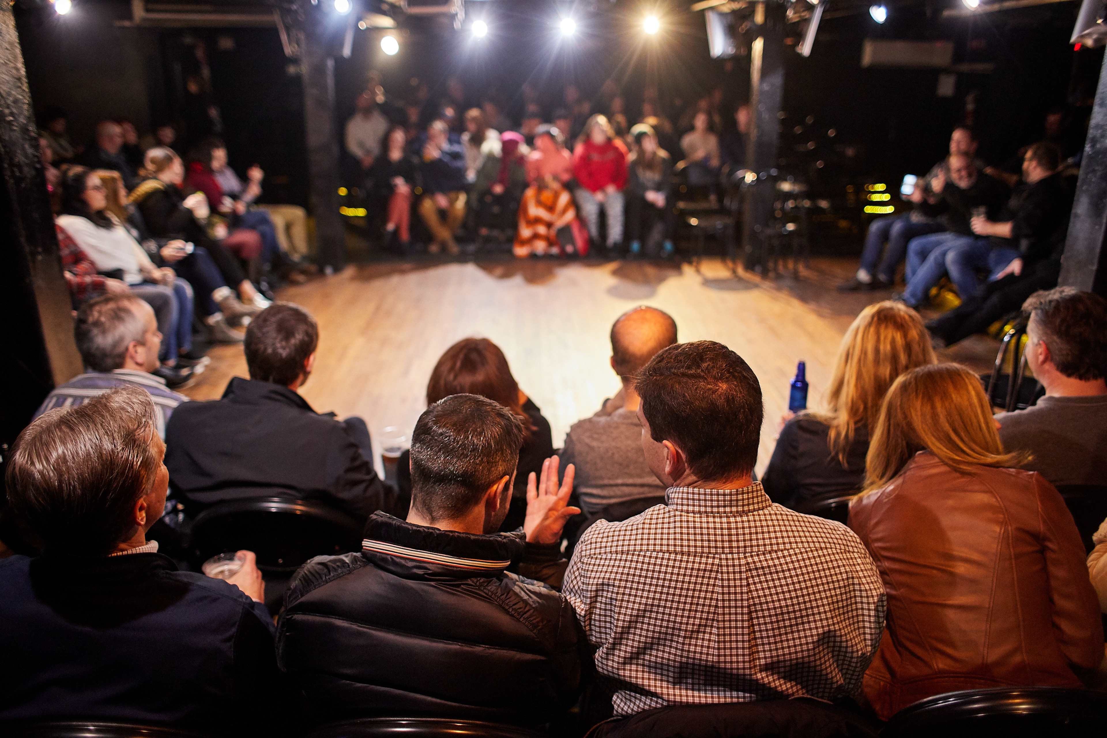 A crowd of spectators sits in a small theater, facing a stage lit by overhead lights.