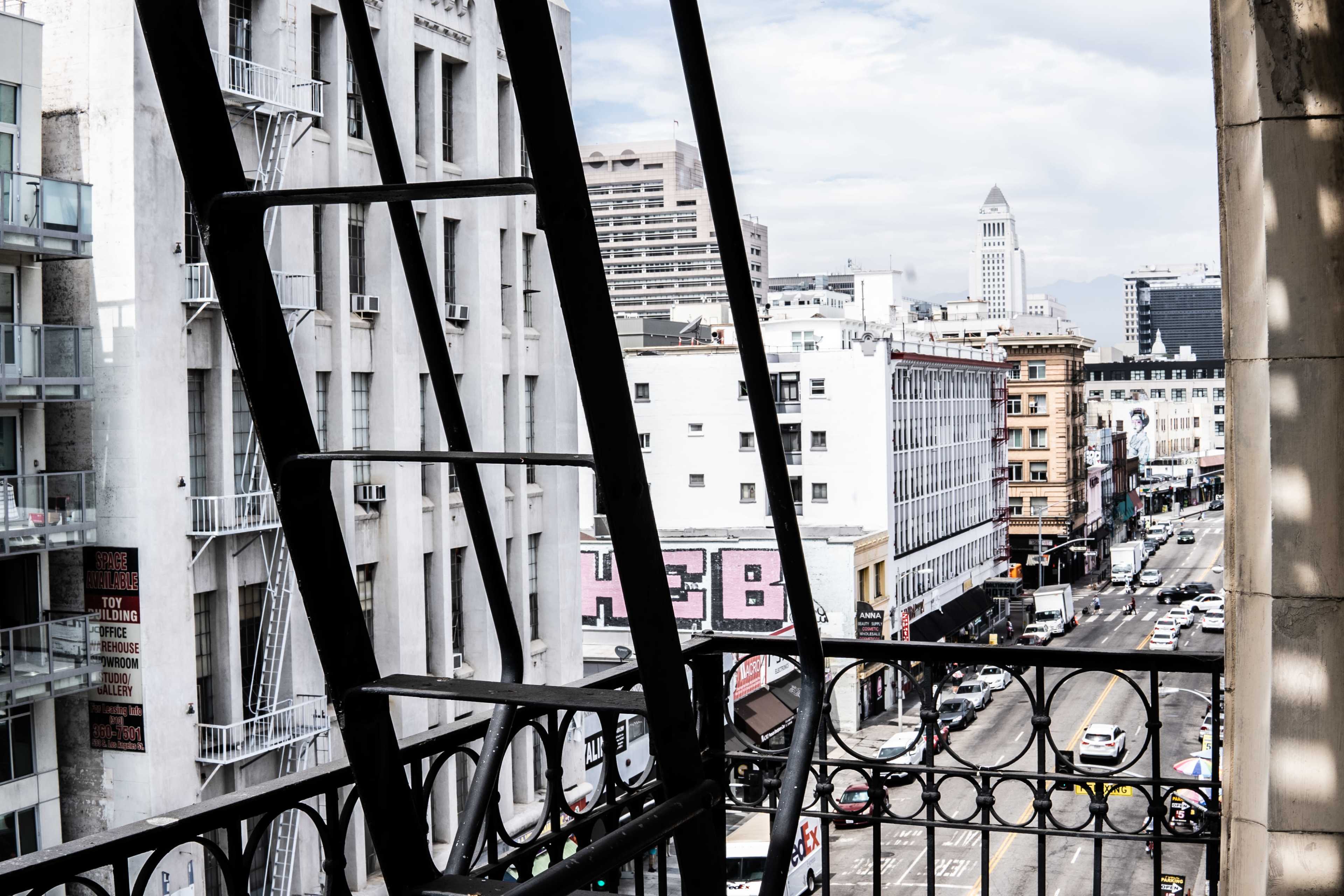 The image shows a view of a city street lined with buildings, framed by an iron fire escape on the left.