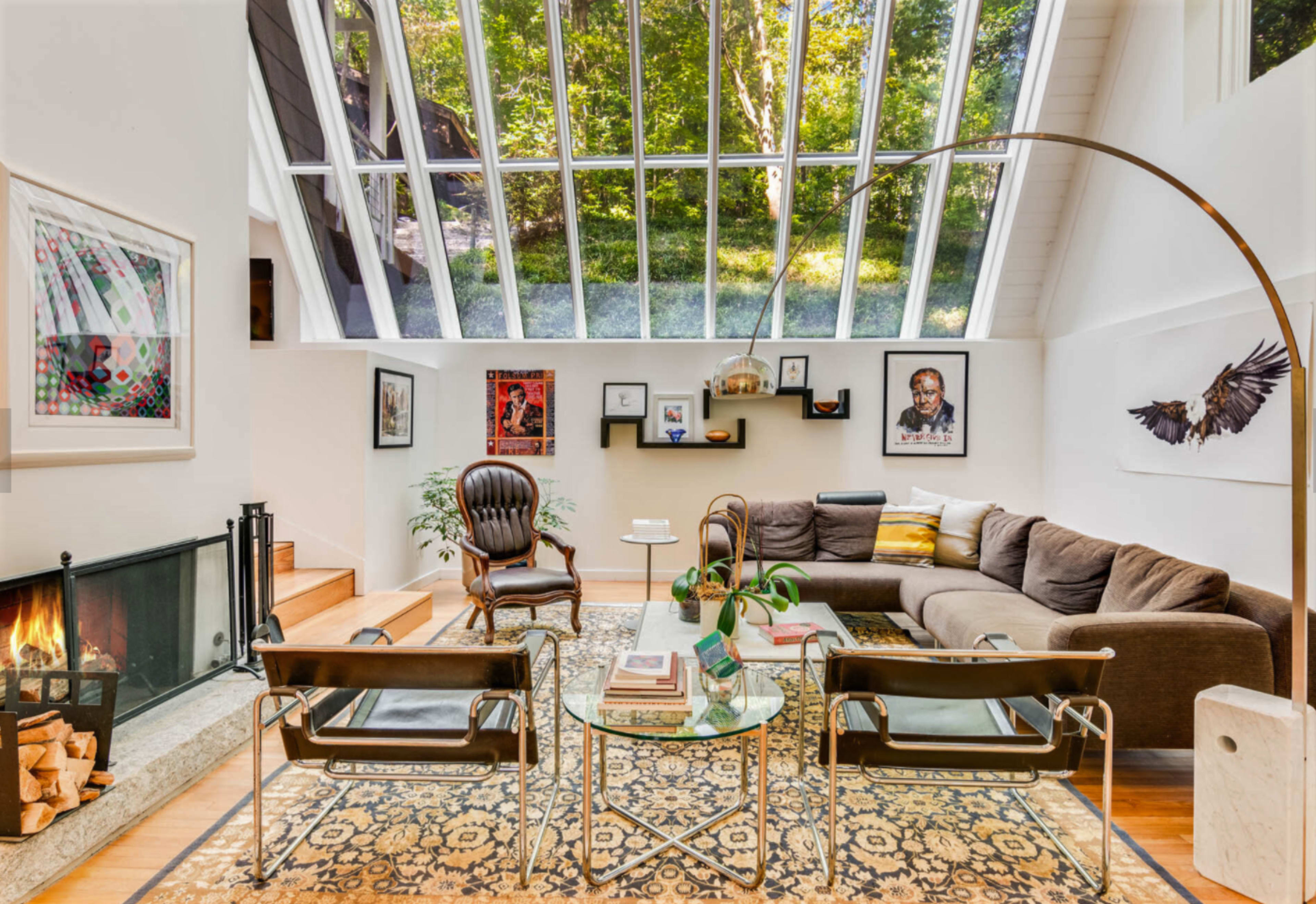 The image shows a modern living room with large glass windows, a sectional sofa, an armchair, a fireplace, and various framed artworks on the walls.