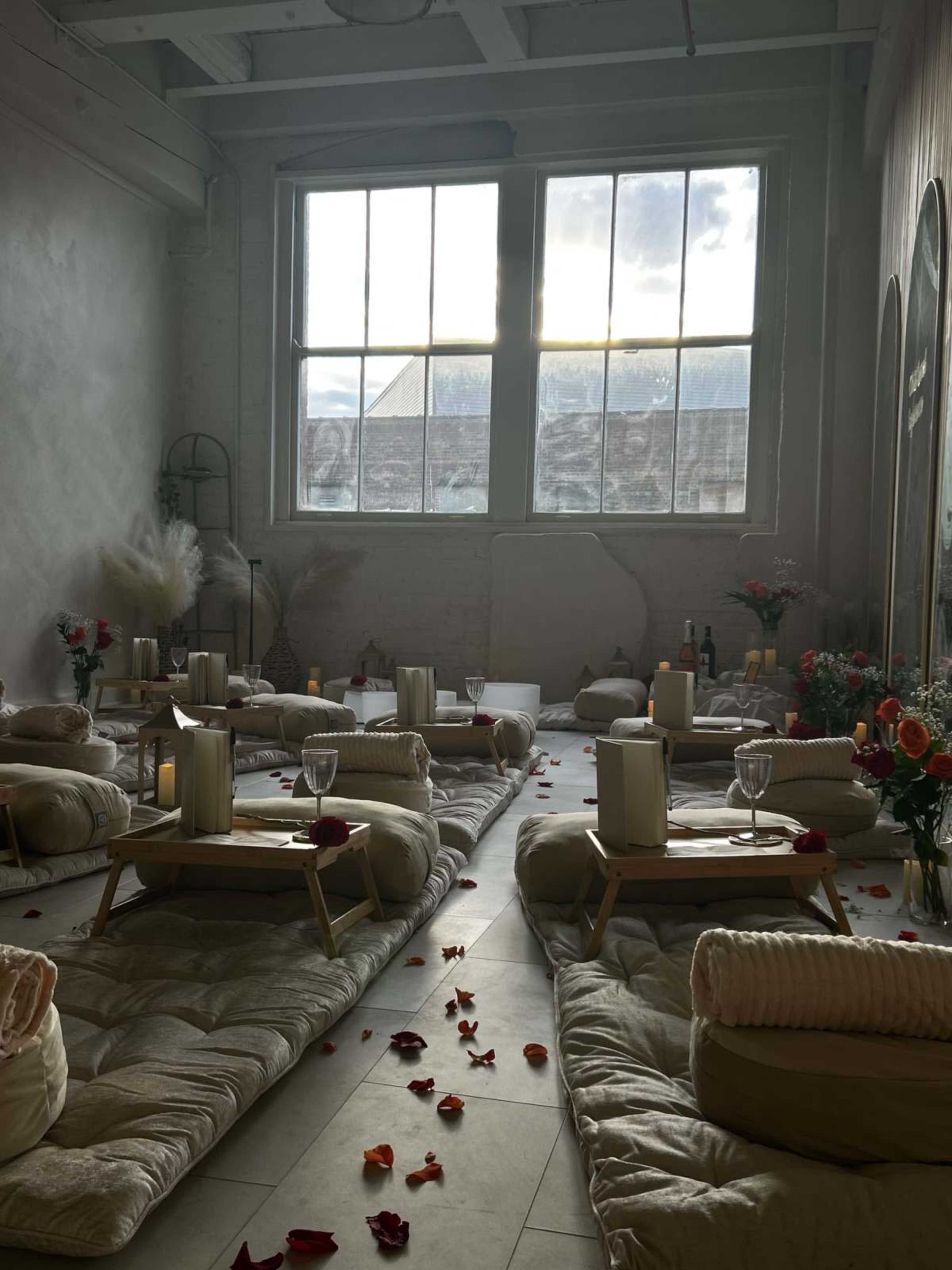 A cozy, softly lit room is arranged with floor cushions and small tables, surrounded by scattered rose petals and flowers, creating an inviting atmosphere.