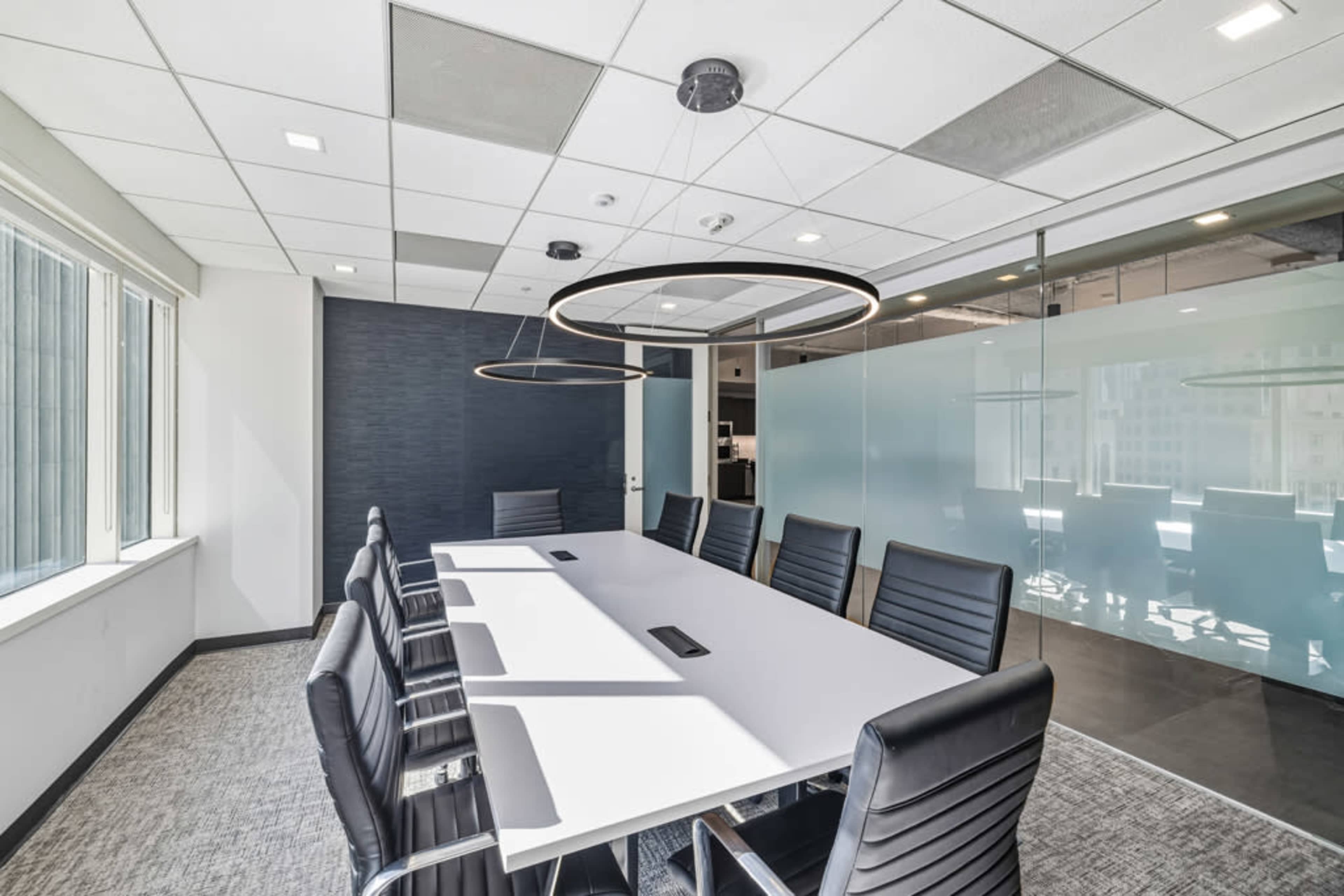 The image shows a modern conference room with a long table surrounded by black chairs, illuminated by circular ceiling lights.