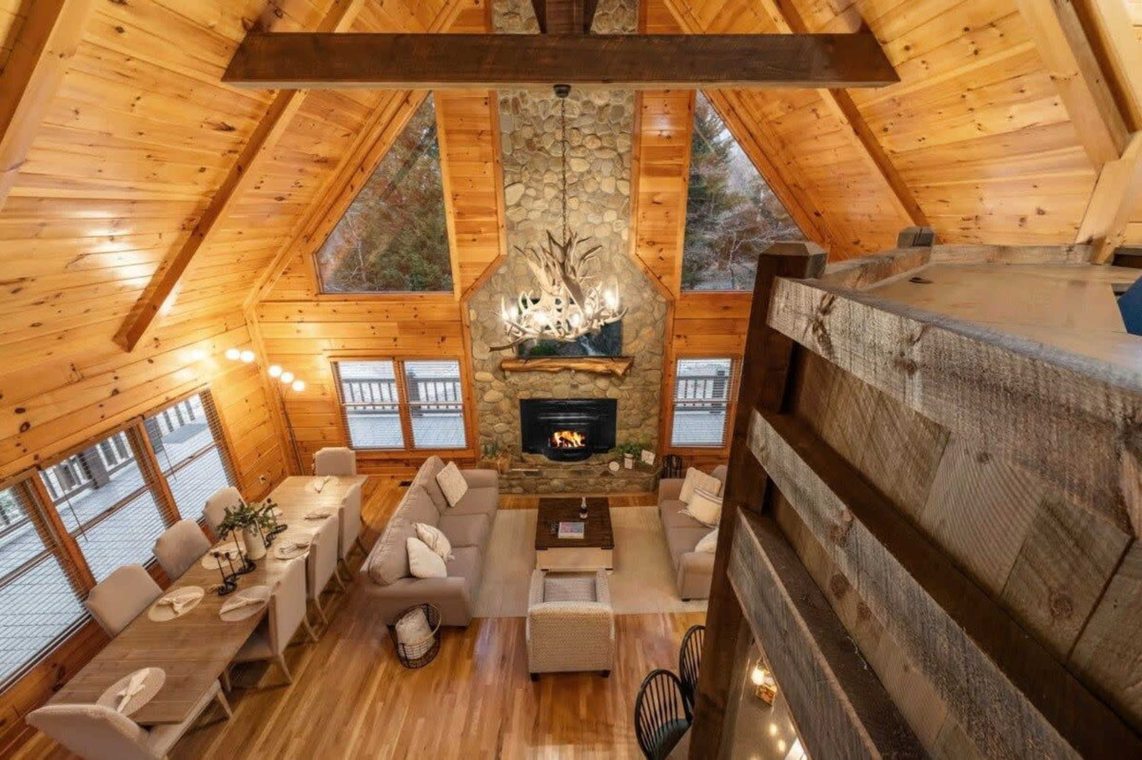 A spacious, rustic cabin interior with wooden walls and a stone fireplace, featuring a dining area and comfortable seating arranged around a central coffee table.