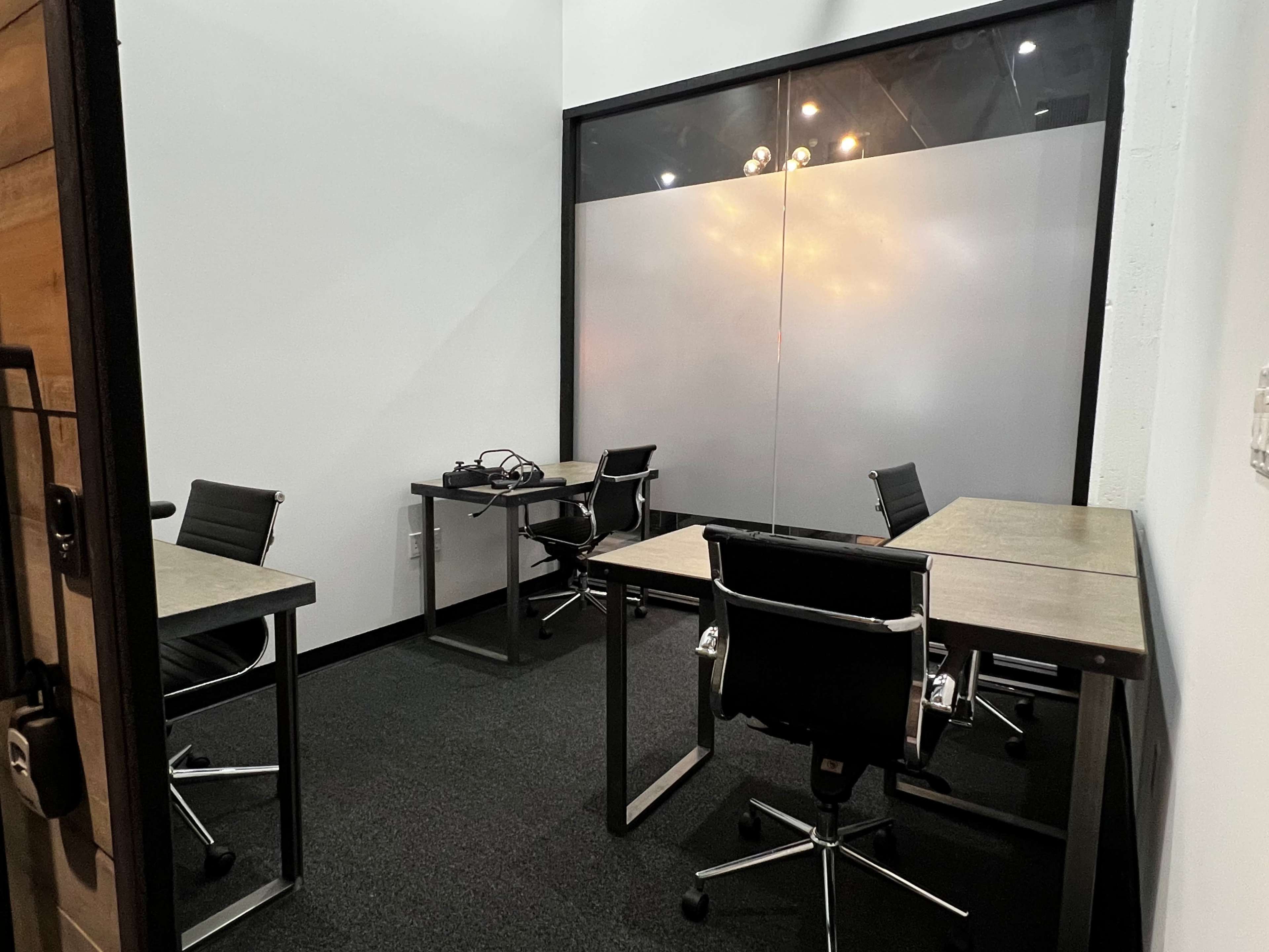 The image shows a small office space featuring two desks with chairs and a glass partition that separates it from the adjacent area.