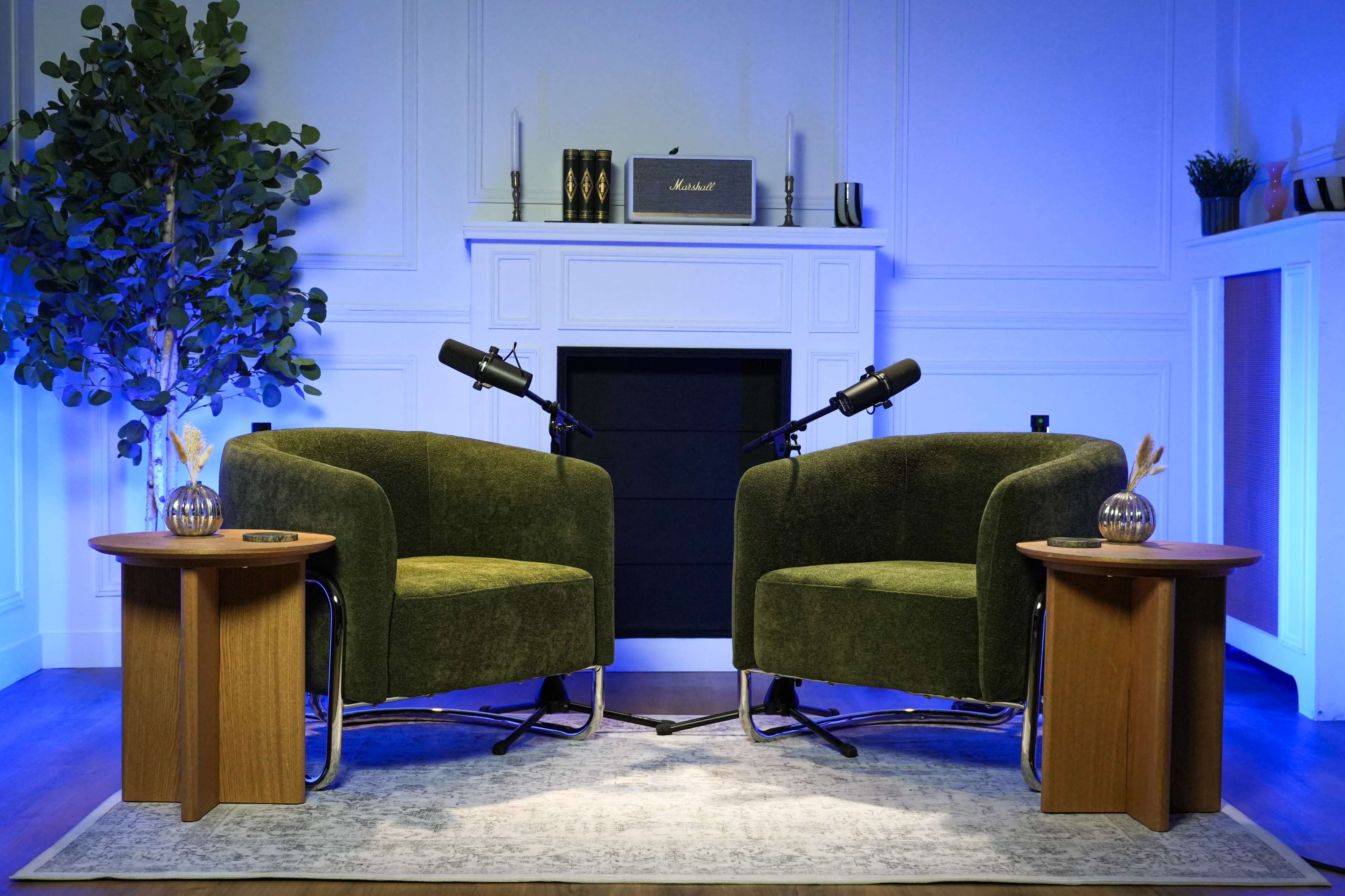 The image features two green armchairs placed facing each other, accompanied by wooden side tables, in a well-lit room with a fireplace and decorative plants.