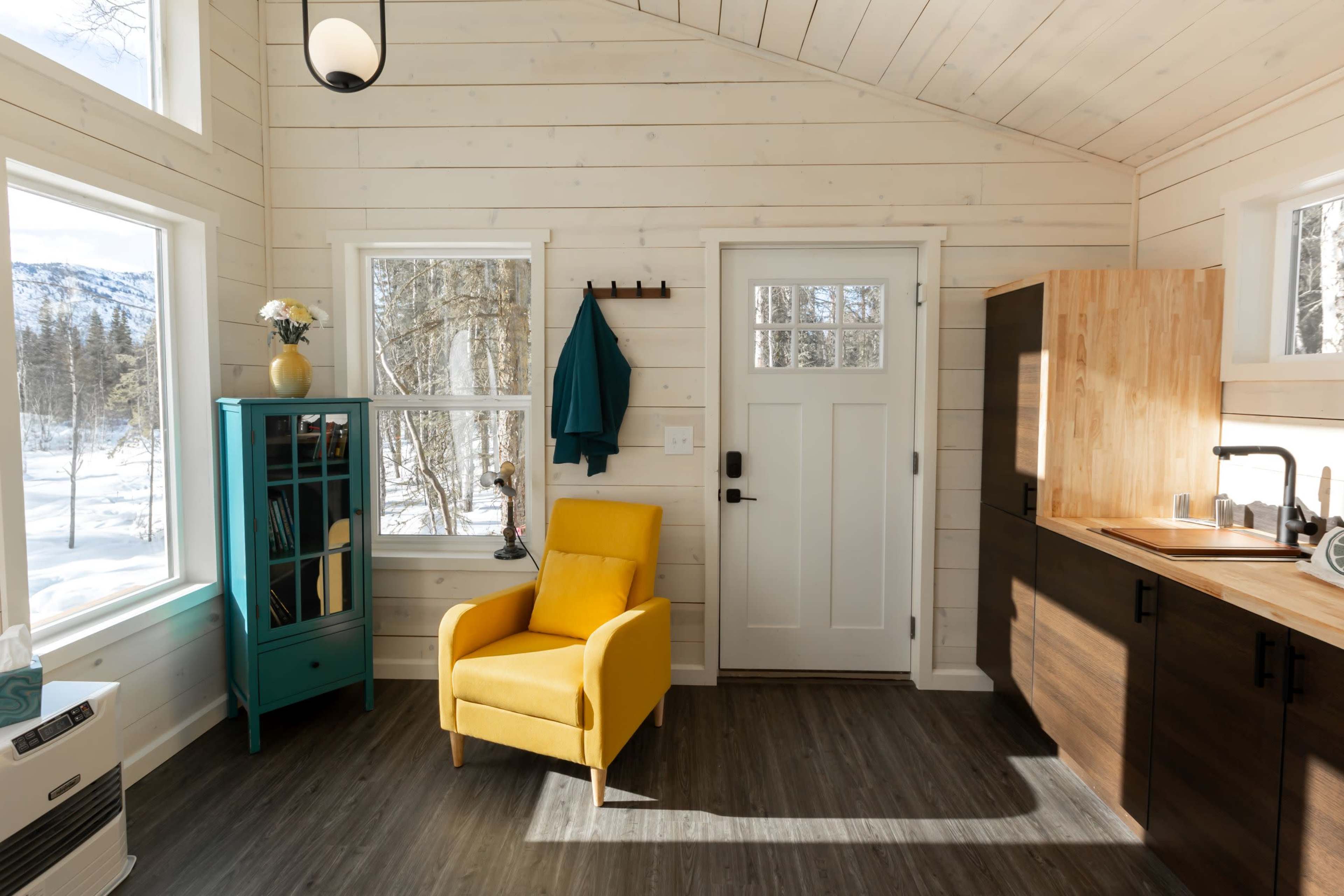 The image shows a small, cozy interior space with a yellow armchair, a teal cabinet, a kitchenette, and large windows overlooking a snowy landscape.