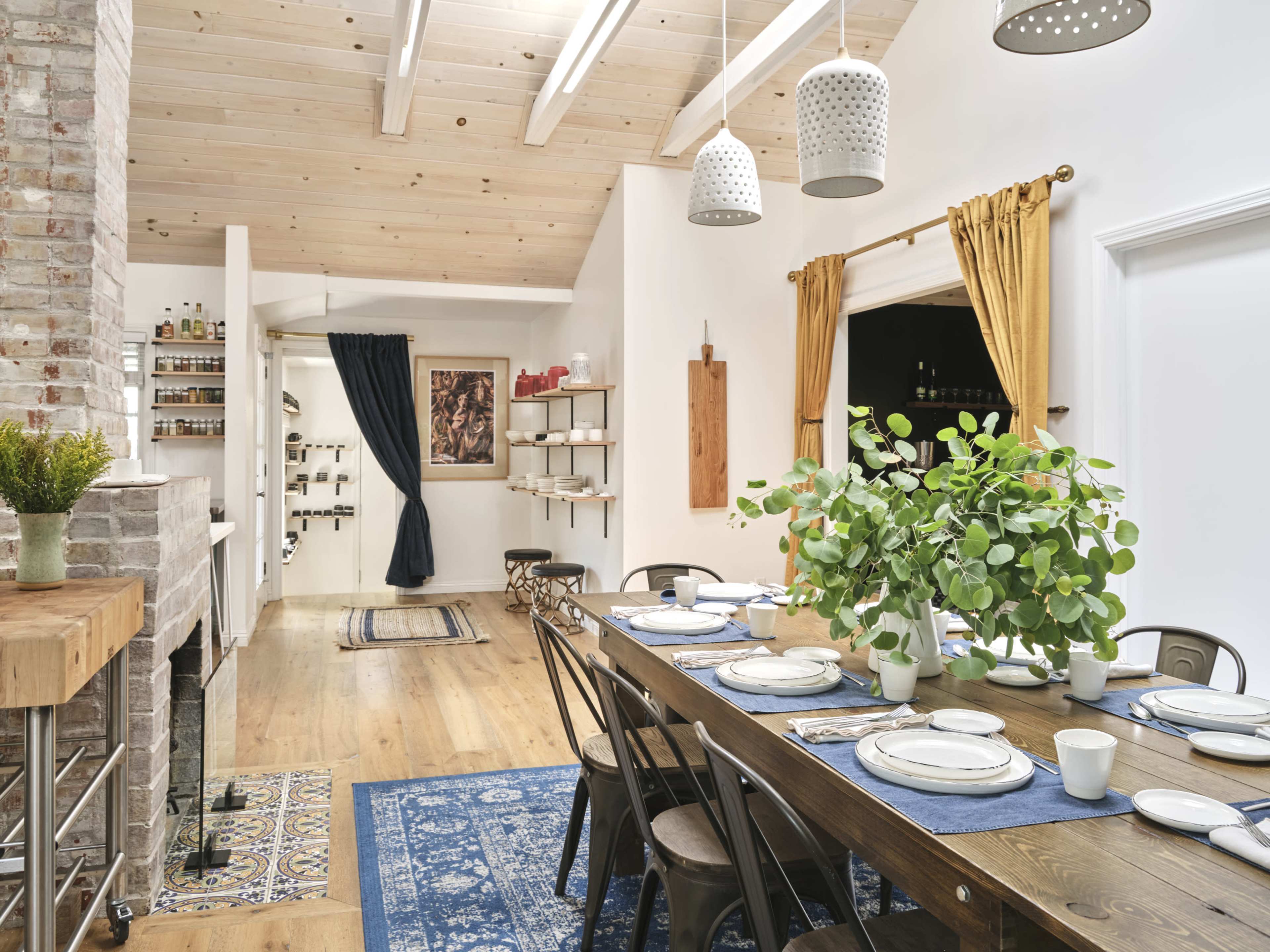 A dining area with a large wooden table set for a meal, surrounded by black chairs and adorned with a leafy centerpiece, against a backdrop of light-colored walls and wooden beams.