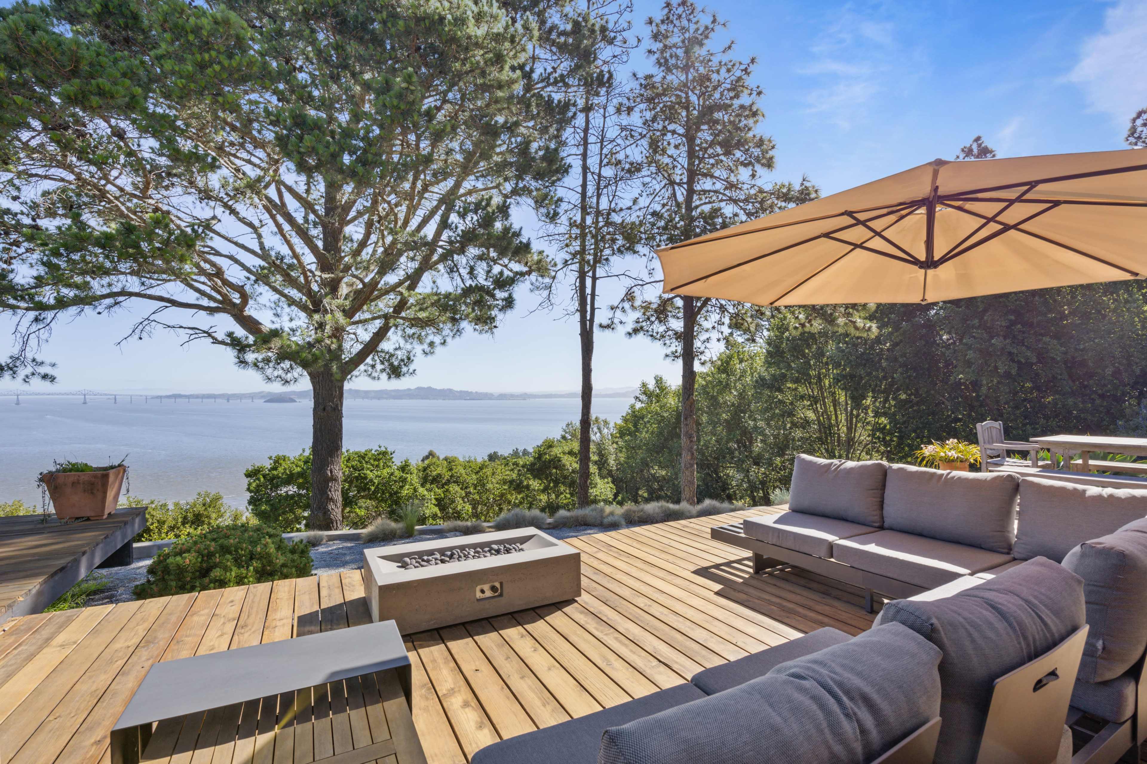 A wooden deck with outdoor seating and a fire pit overlooking a body of water and trees.