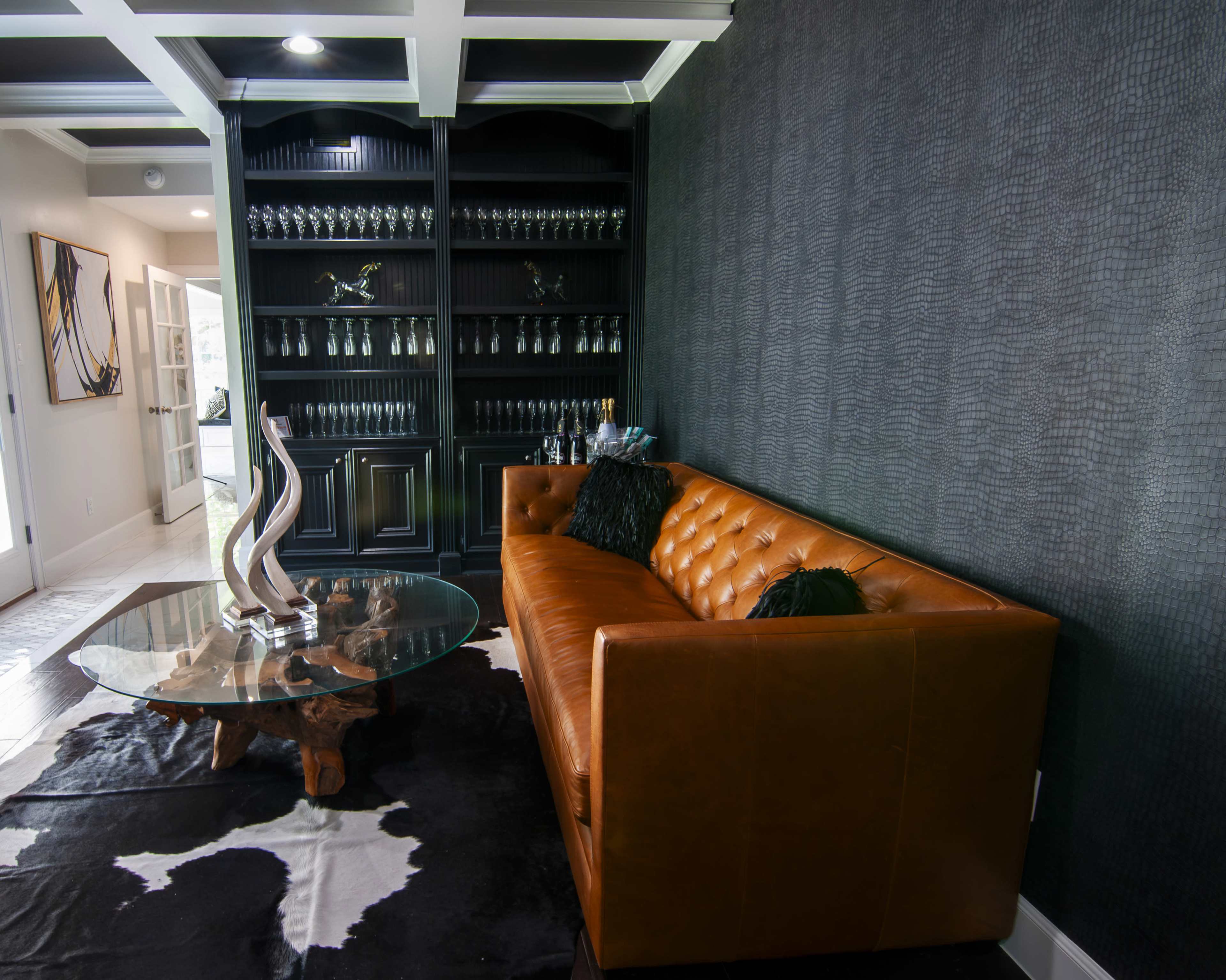 A modern living room features a brown leather couch, a glass coffee table, and a black wall with shelves displaying glassware.