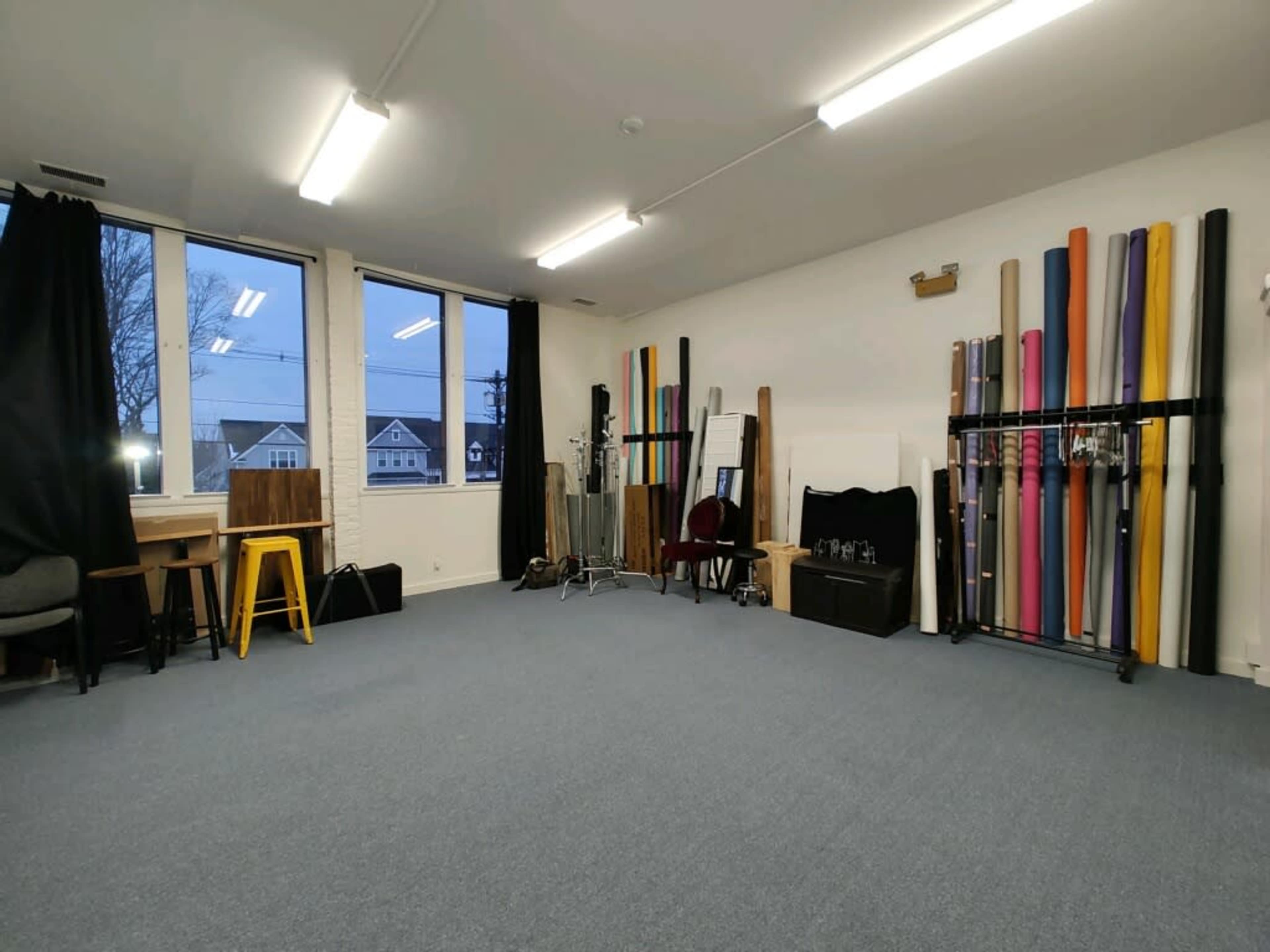 A spacious interior studio with a carpeted floor, large windows, and various rolls of colored backdrops neatly arranged against the wall.