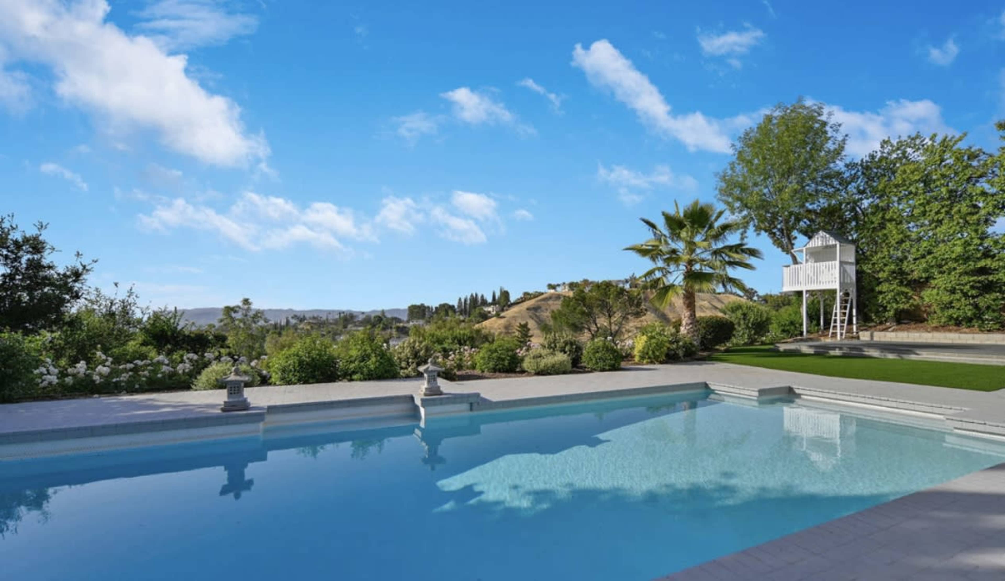 A clear blue swimming pool surrounded by green landscaping and a distant hill under a partly cloudy sky.
