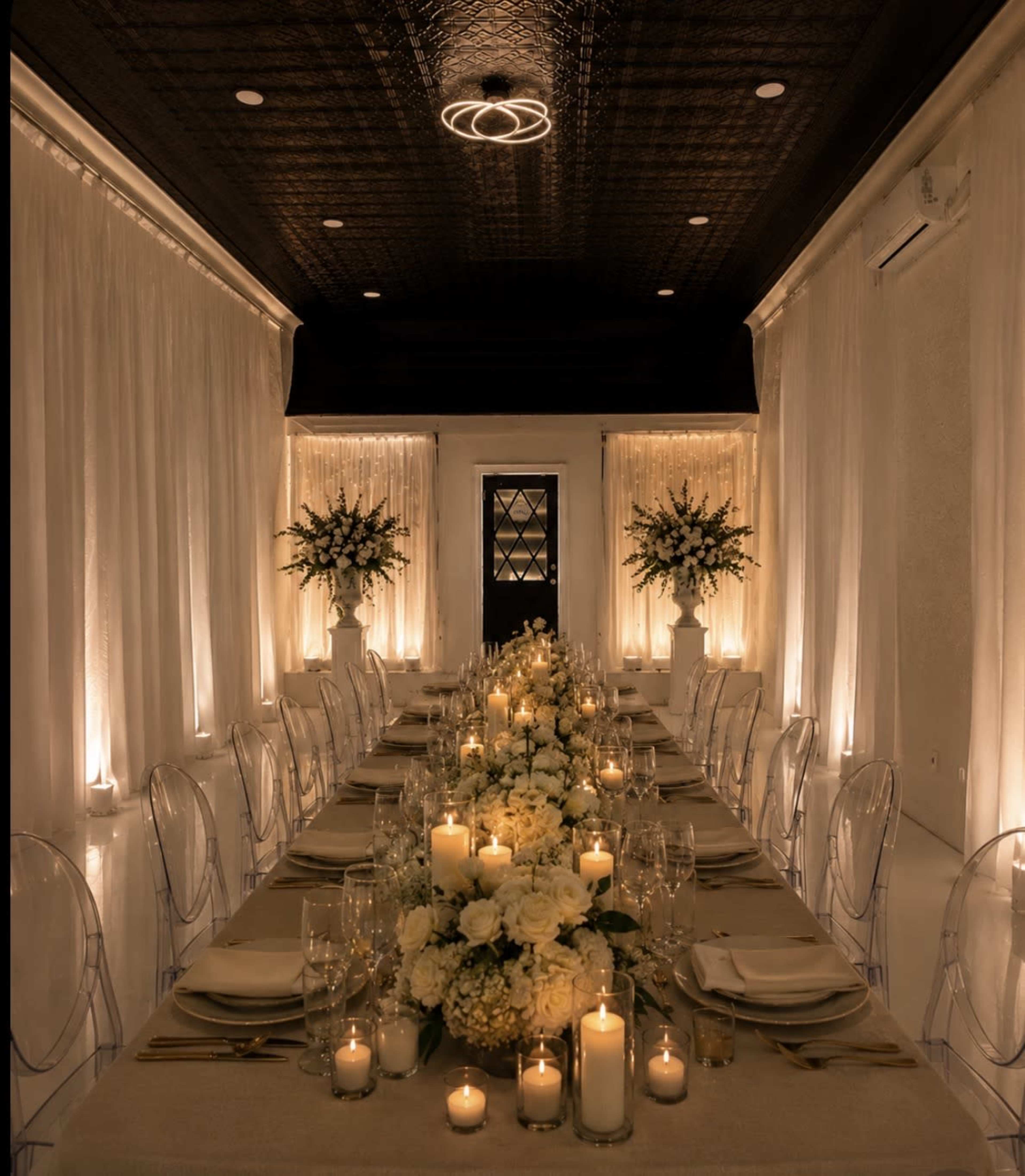 A long dining table is set with candles and floral arrangements in a softly lit, elegant room with white drapes.