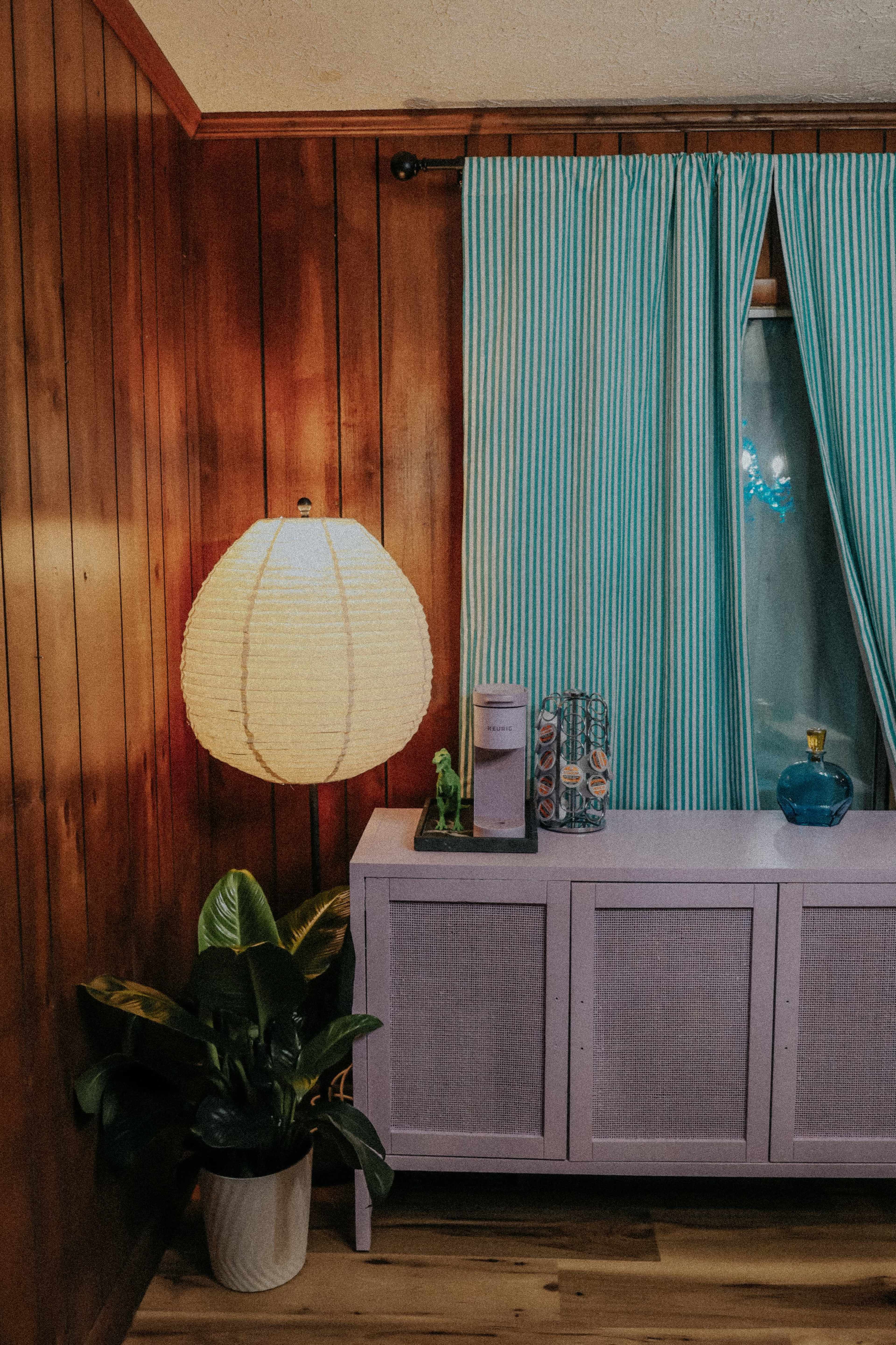 A purple sideboard with a white lamp, blue decorative items, and striped curtains is placed against a wooden wall with a potted plant in the corner.