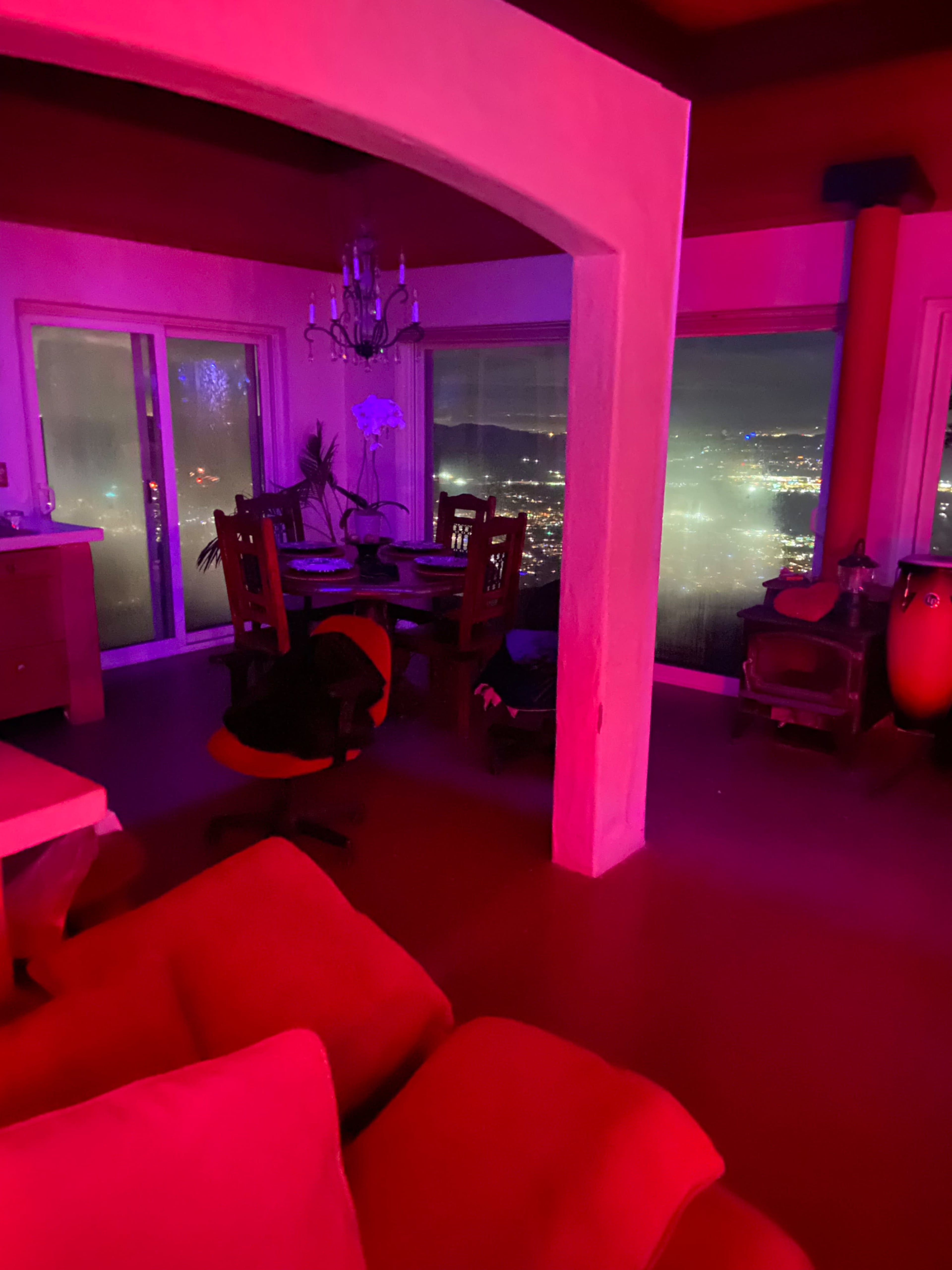 The interior of a dimly lit room features red ambient lighting, a dining area with chairs and a table, and a view of a cityscape through large windows.