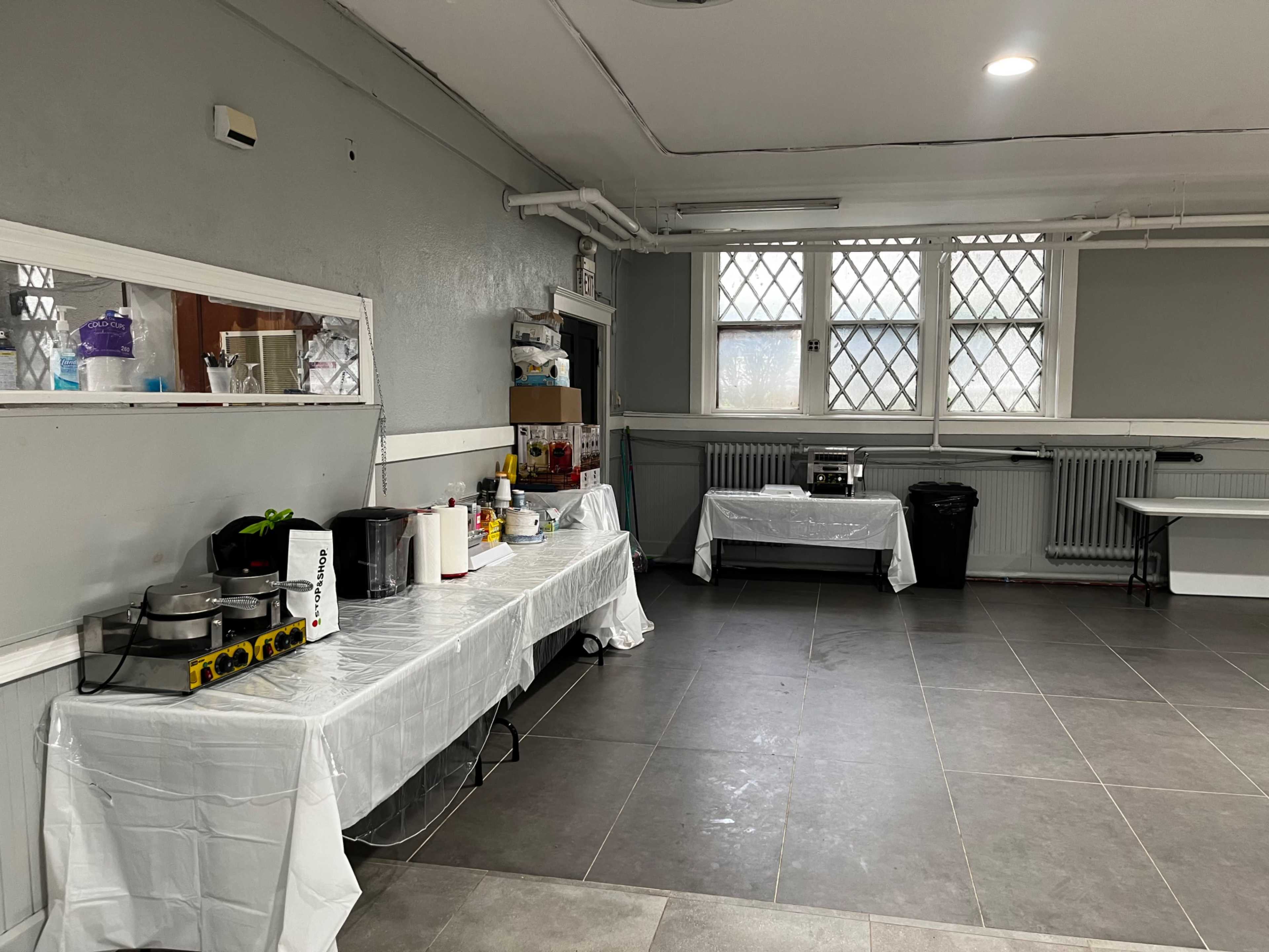 A clean kitchen space with multiple tables covered in white tablecloths, appliances, and a large window with metal grilles.