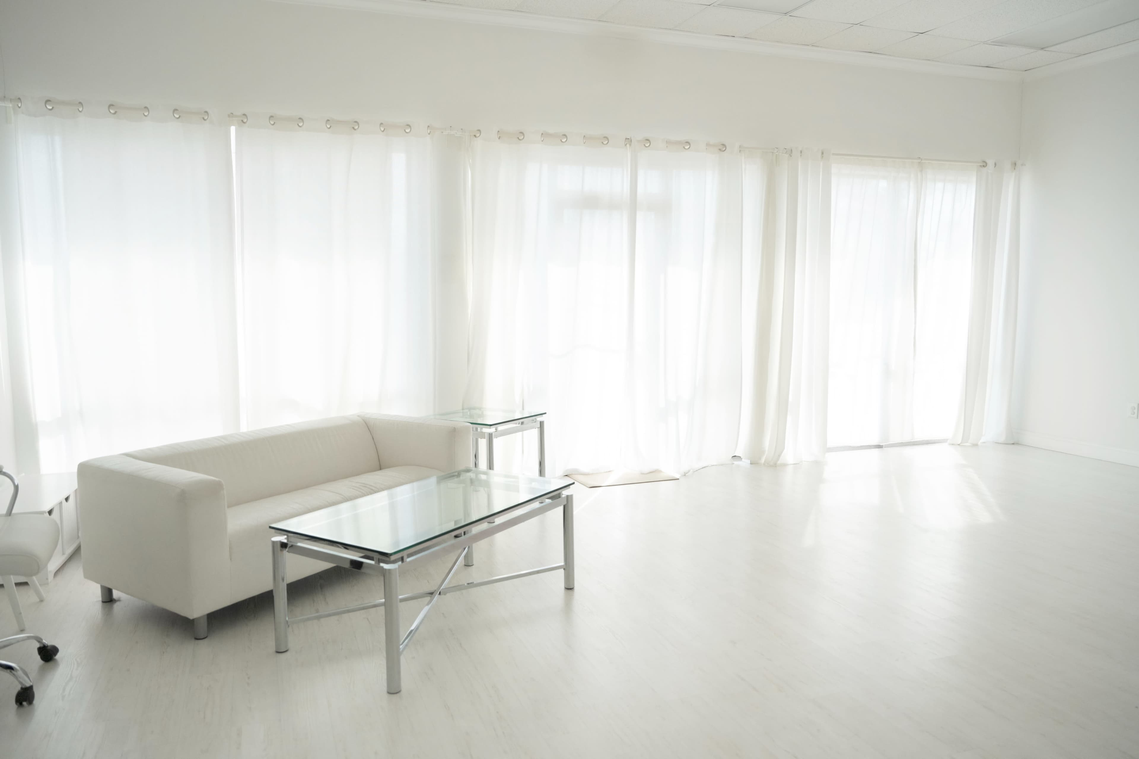 The image shows a bright, spacious room featuring a white sofa, a glass coffee table, and large windows with sheer curtains.