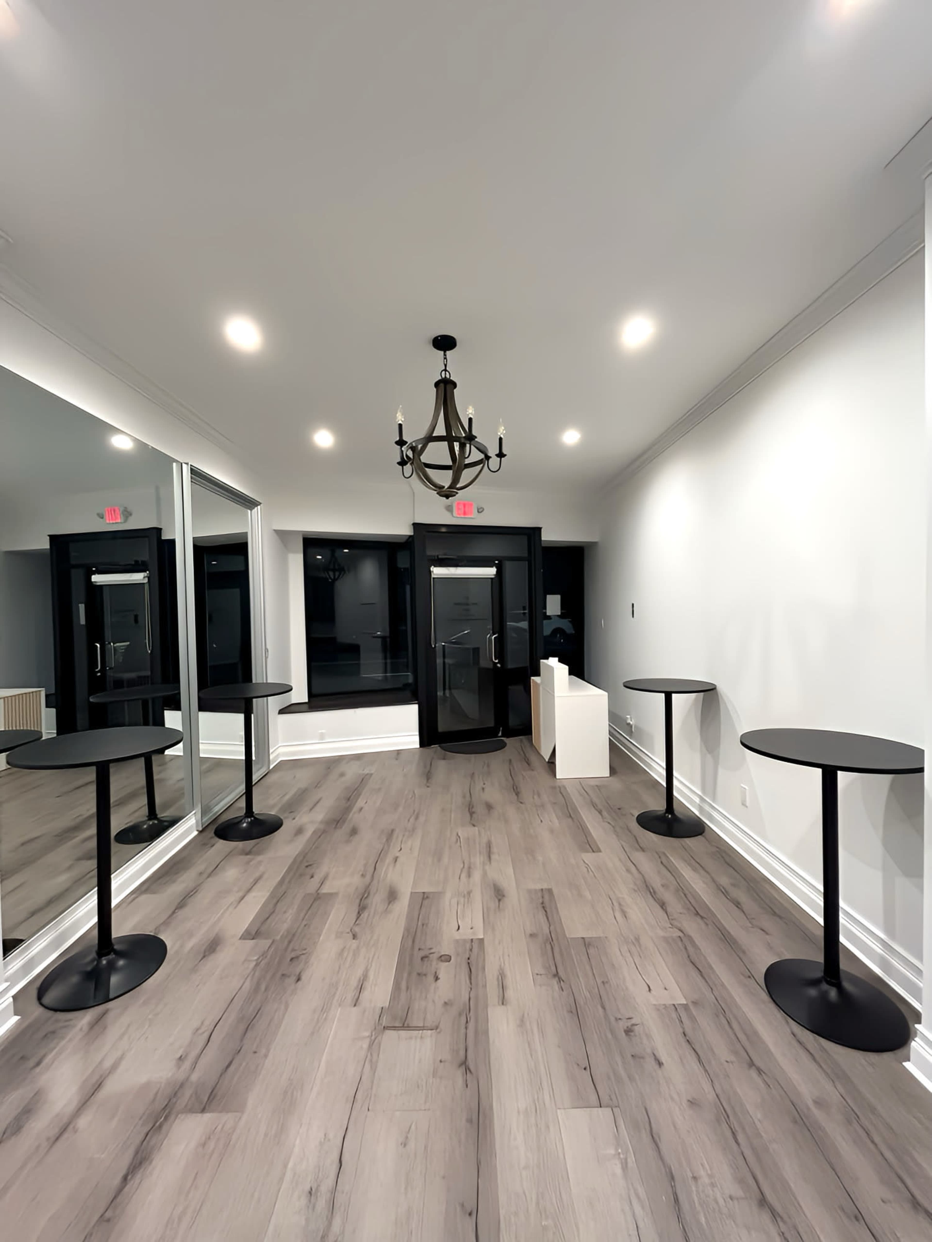 The image shows a well-lit room with wooden flooring, featuring three tall, round tables and a large mirror along one wall, leading to a glass entrance door.