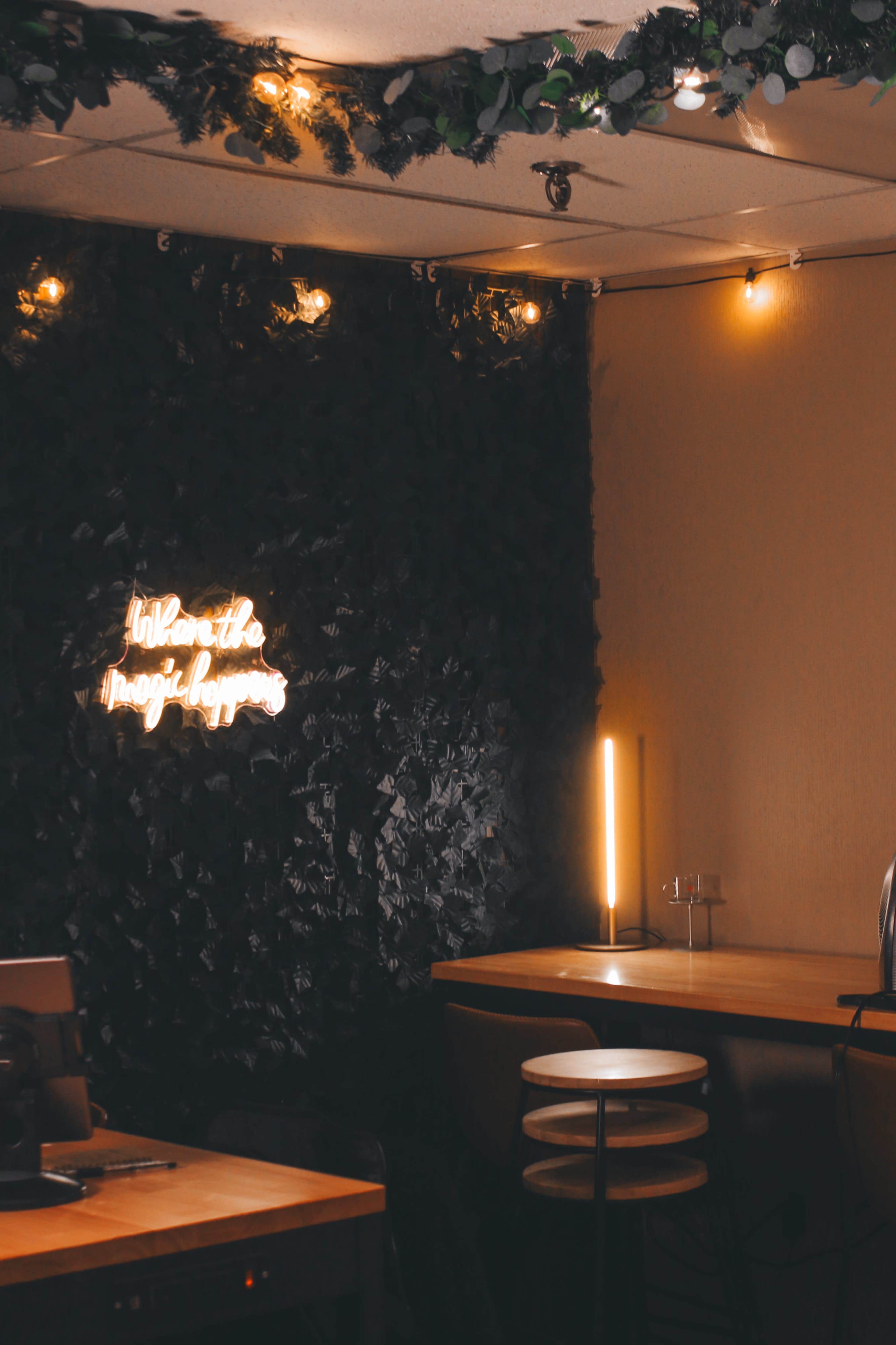 The image shows a cozy interior space with a dark, leafy wall, warm lighting, and a wooden table accompanied by stools.
