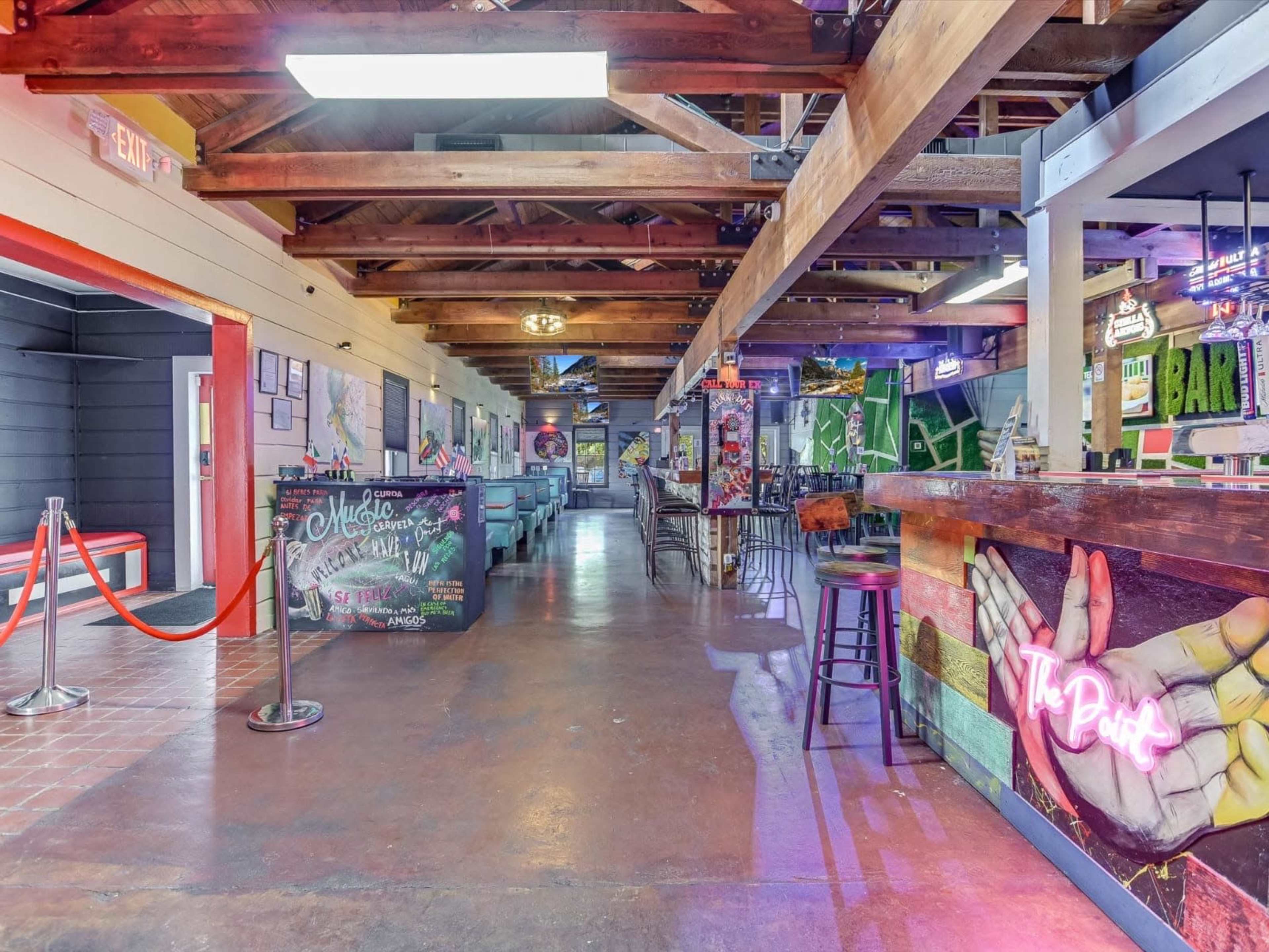 The image shows the interior of a colorful bar with wooden beams, tables, and a long counter displaying neon signage.