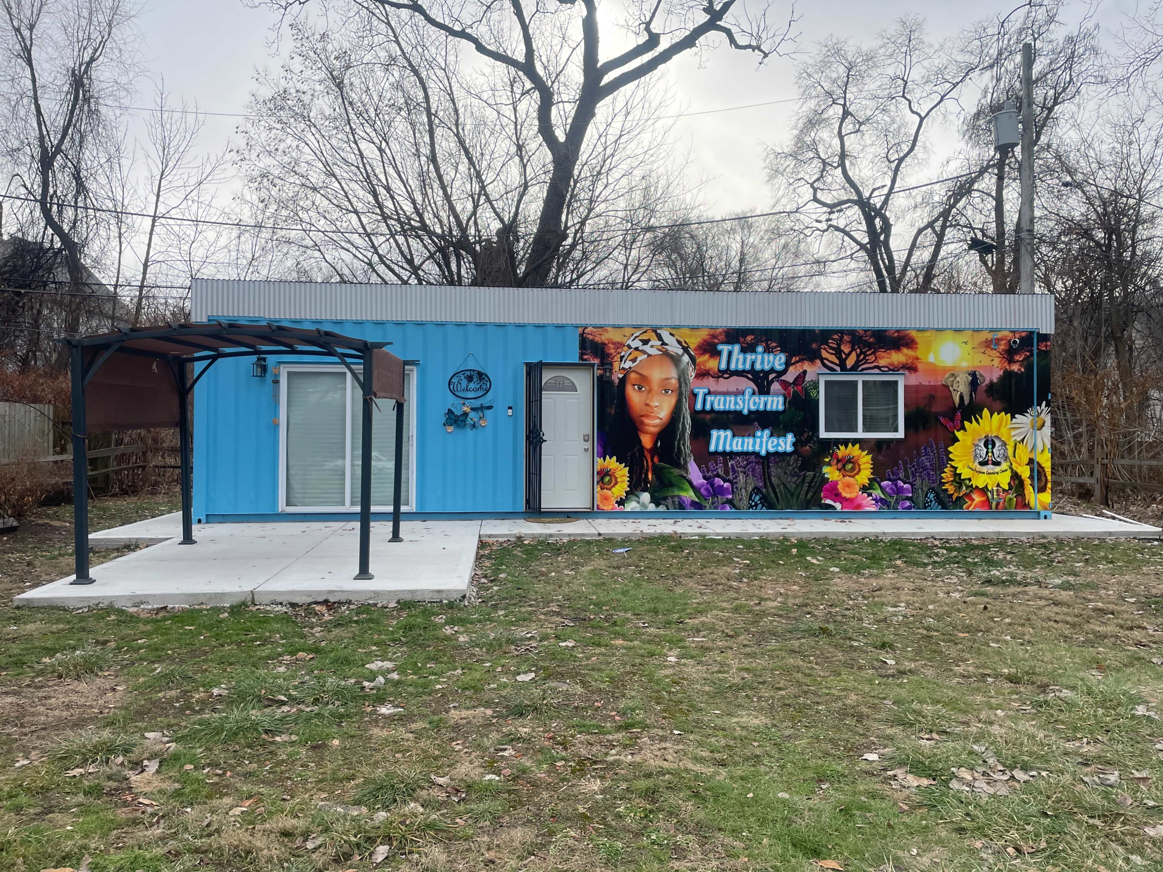 The image shows a brightly painted blue container home with a mural featuring flowers and a woman's face on one side, set in a bare outdoor area.