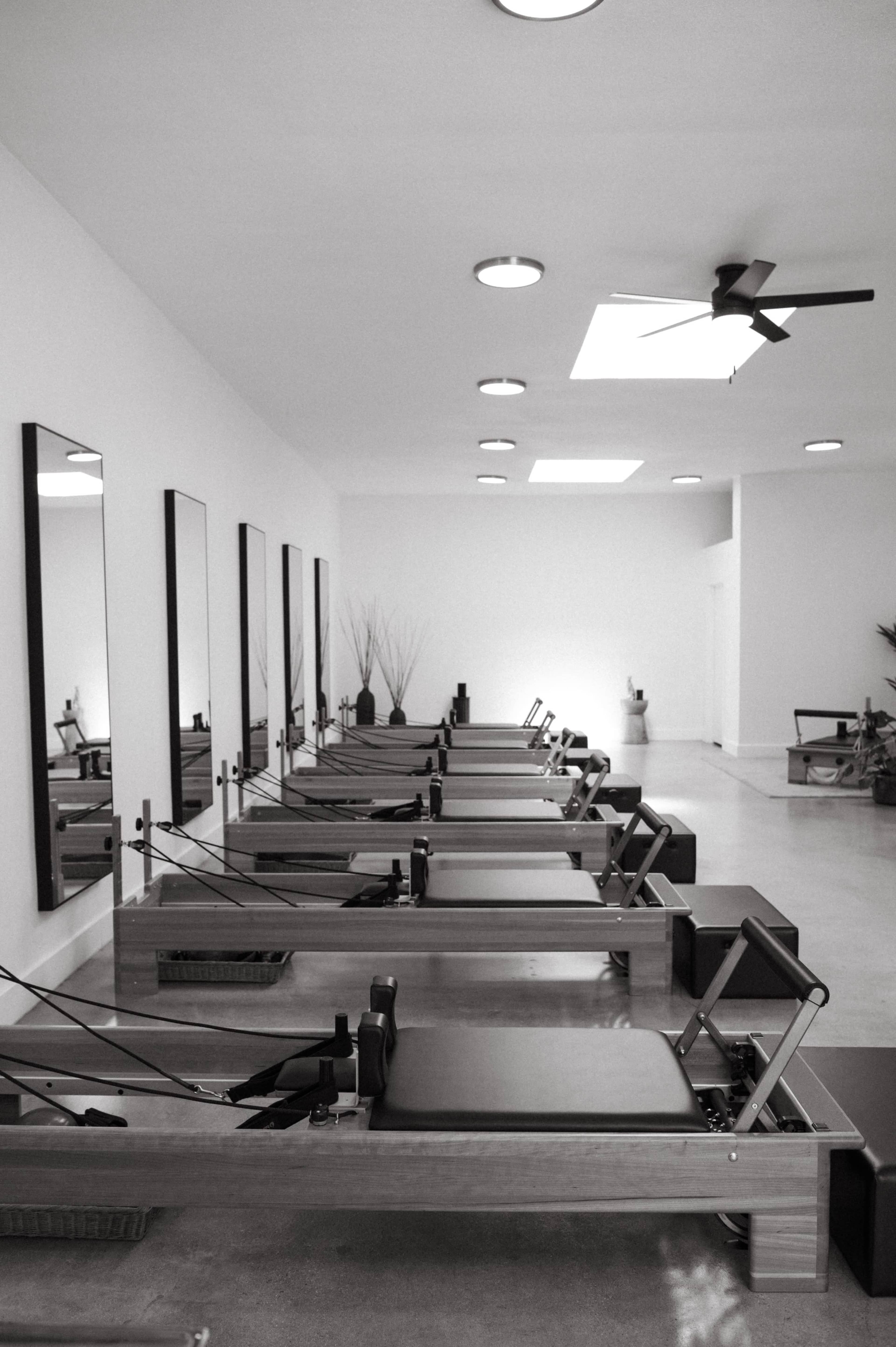 The image shows a row of Pilates reformer machines in a bright, minimalist studio with mirrors on the walls.