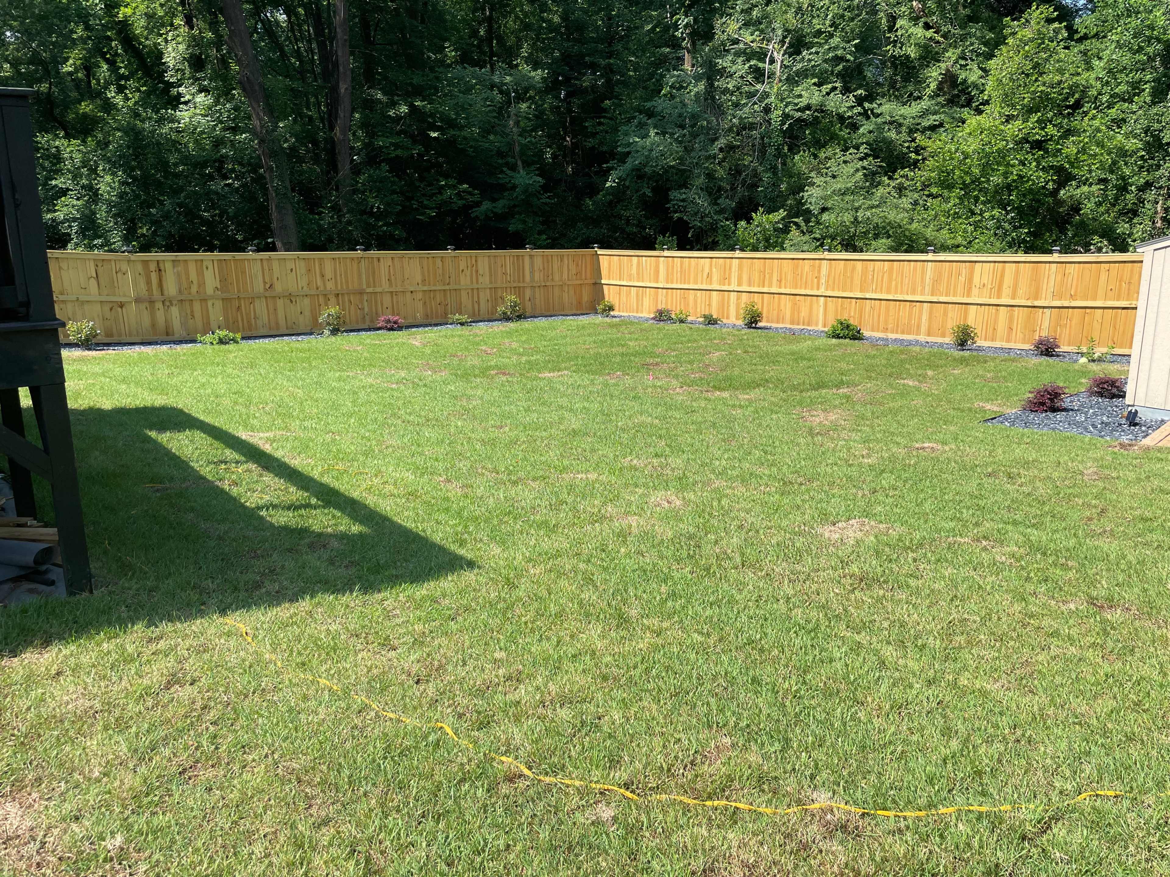 The image shows a grassy yard enclosed by a wooden fence, with landscaping along the perimeter.