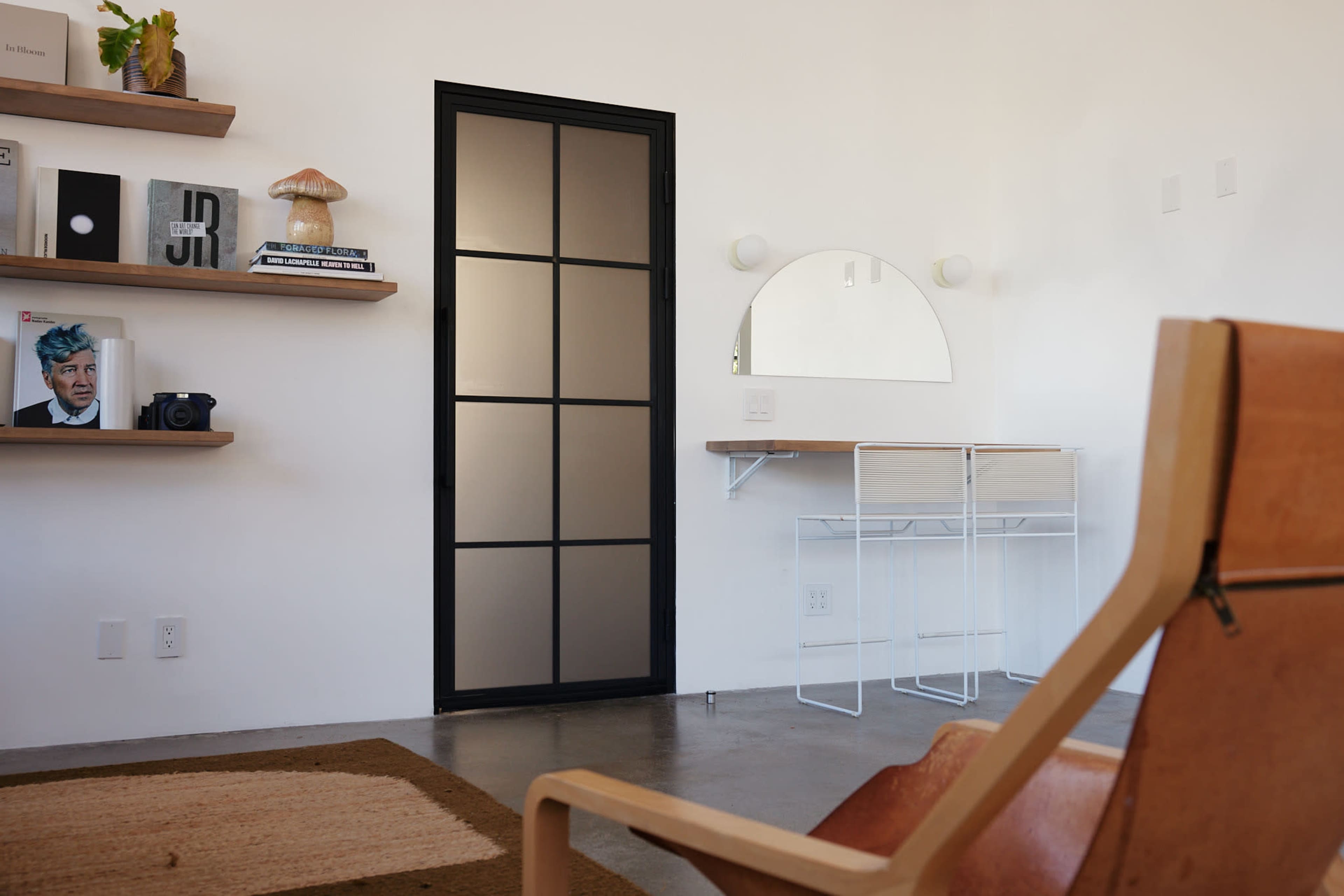 A minimalist interior features a sleek black door, a round mirror, a small desk, and shelves displaying decorative items and a camera.