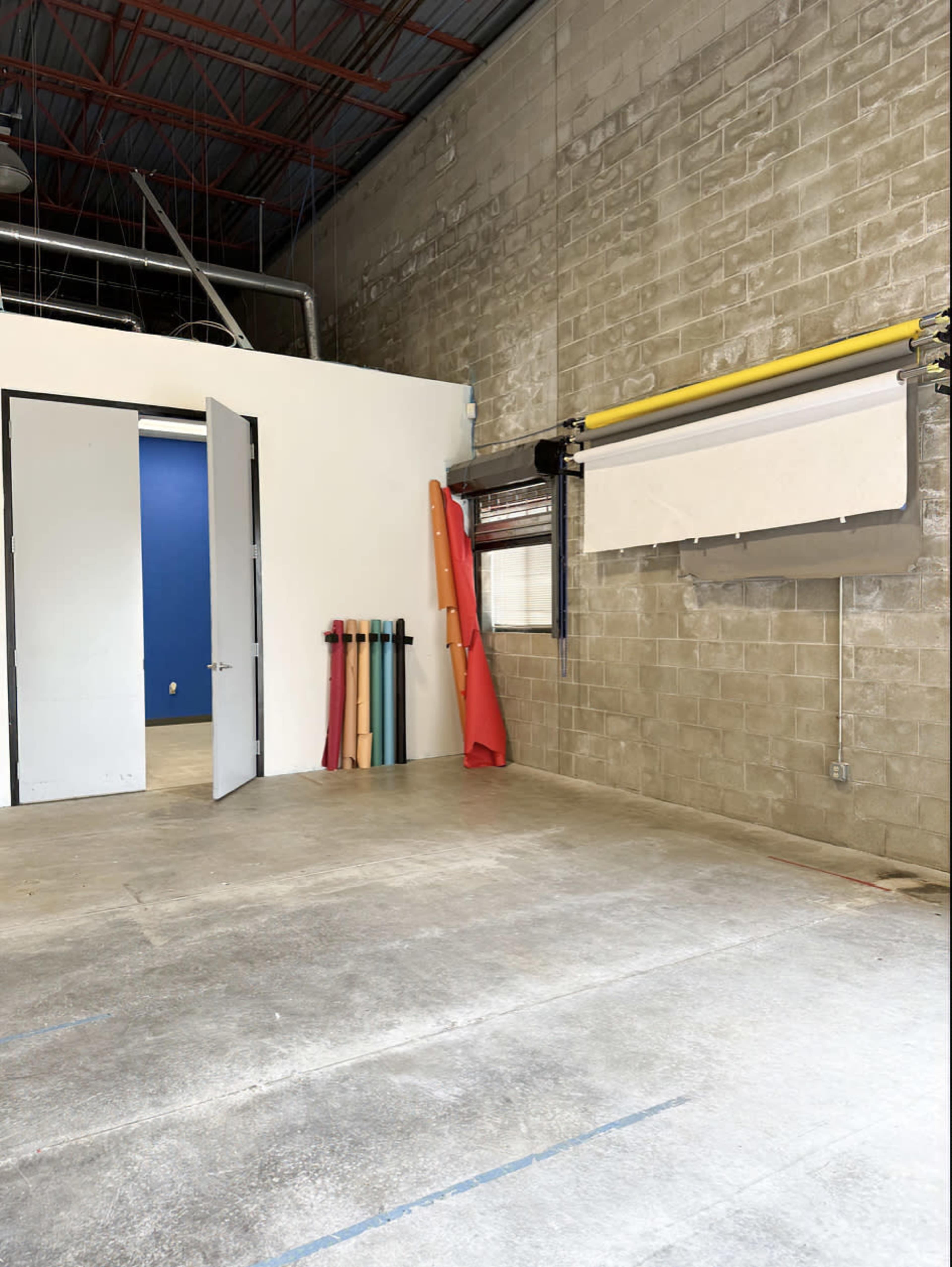 The image shows an industrial space with a gray concrete floor, a blue wall on the left, two open doors, and rolls of colored material leaning against the wall.