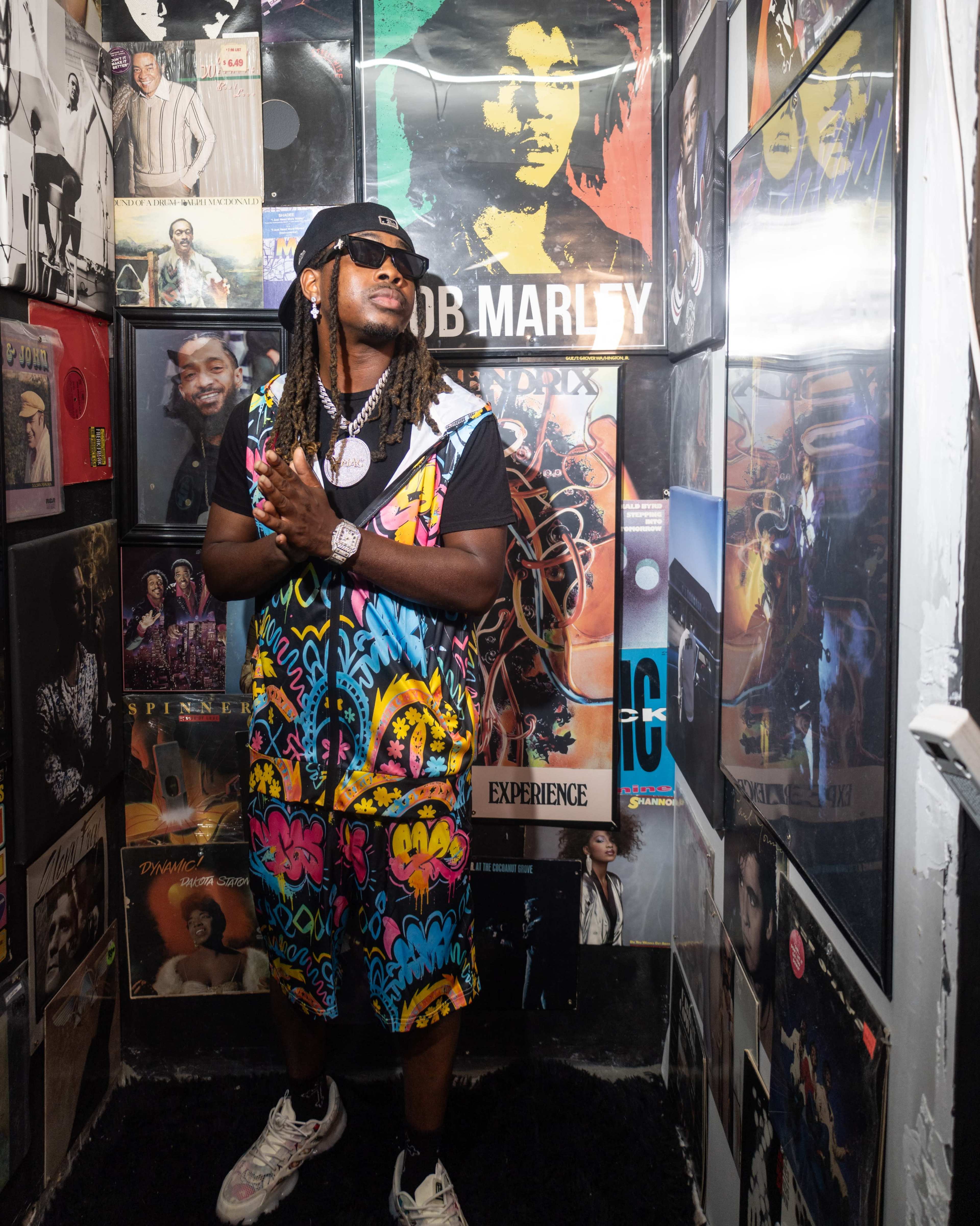A man wearing a colorful outfit stands in a narrow hallway lined with framed music album covers.
