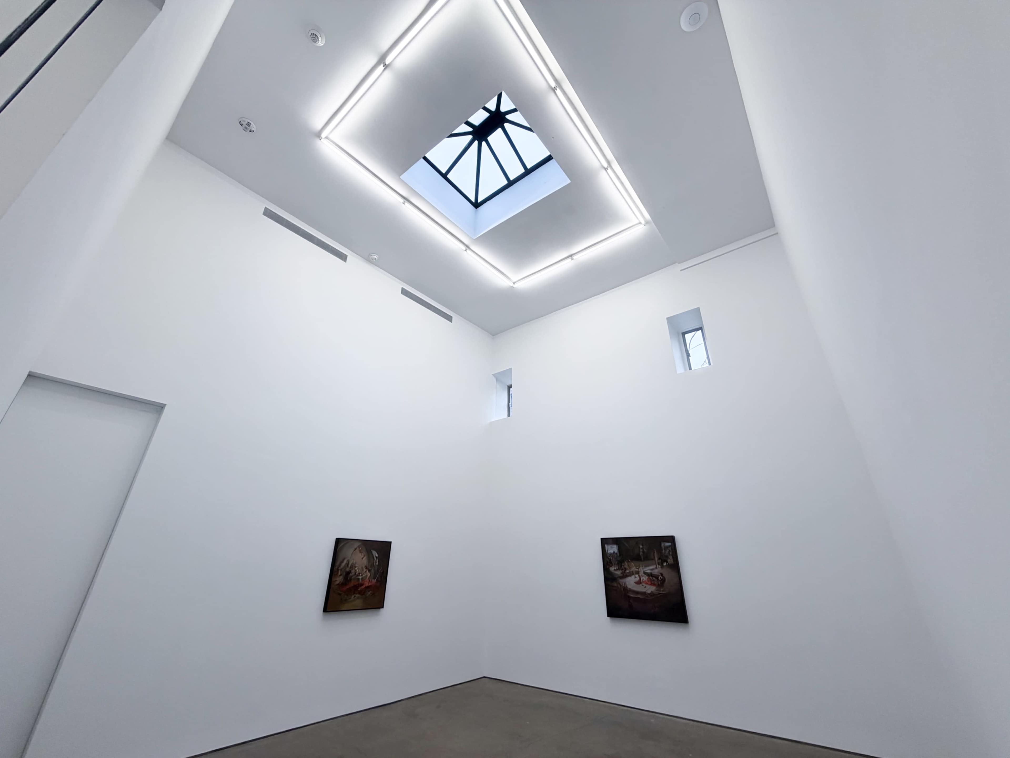 The image shows a minimalist gallery space with white walls, two small windows, and a skylight, featuring two framed paintings on the wall.