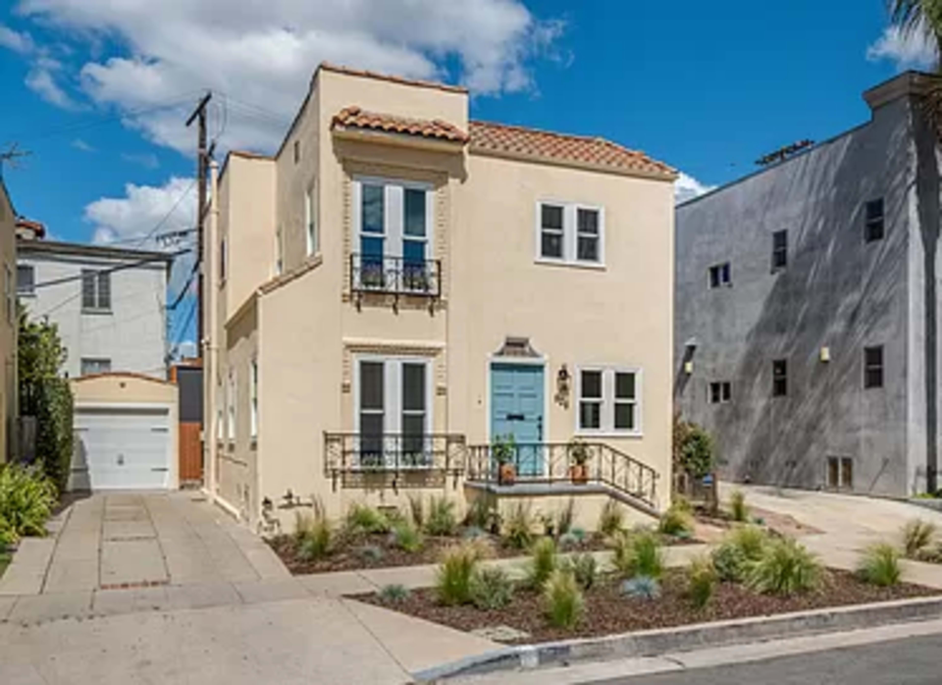 A two-story, light-colored house with a tile roof and blue front door is situated on a landscaped corner lot.