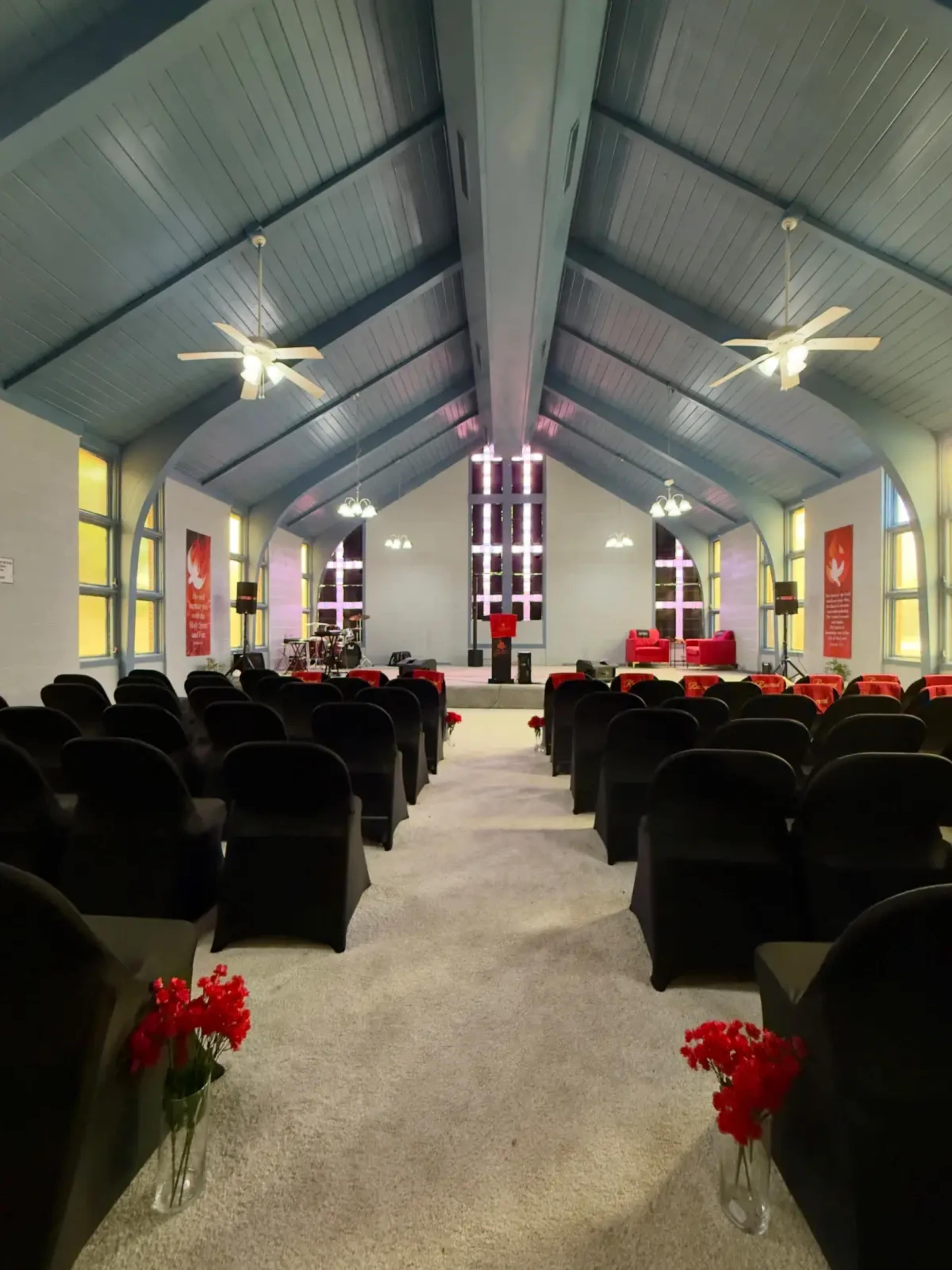 The interior of a church features rows of black chairs, a stage with a cross, and colorful stained glass windows.