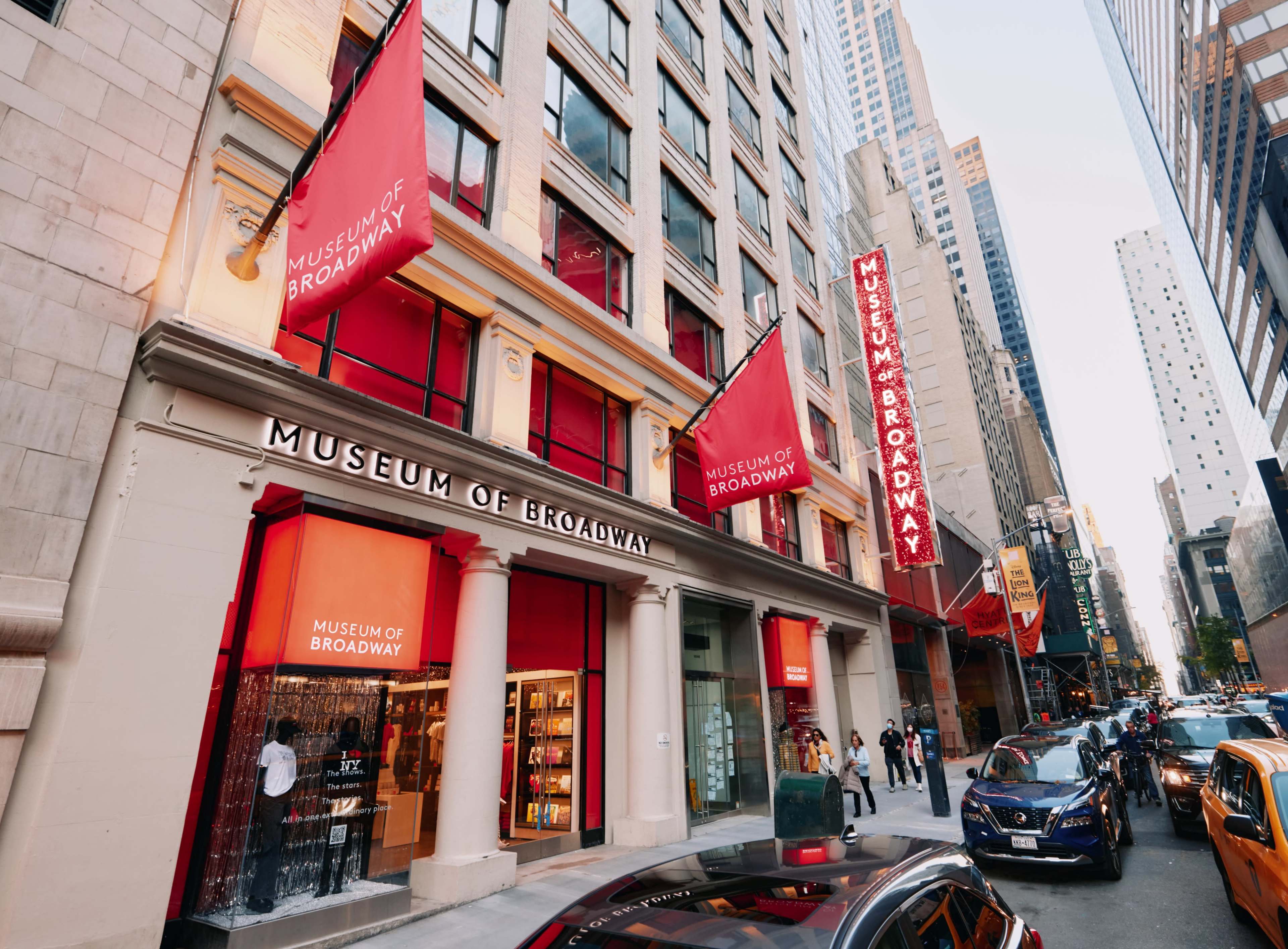 The exterior of the Museum of Broadway is shown on a city street, featuring large red banners with its name prominently displayed.