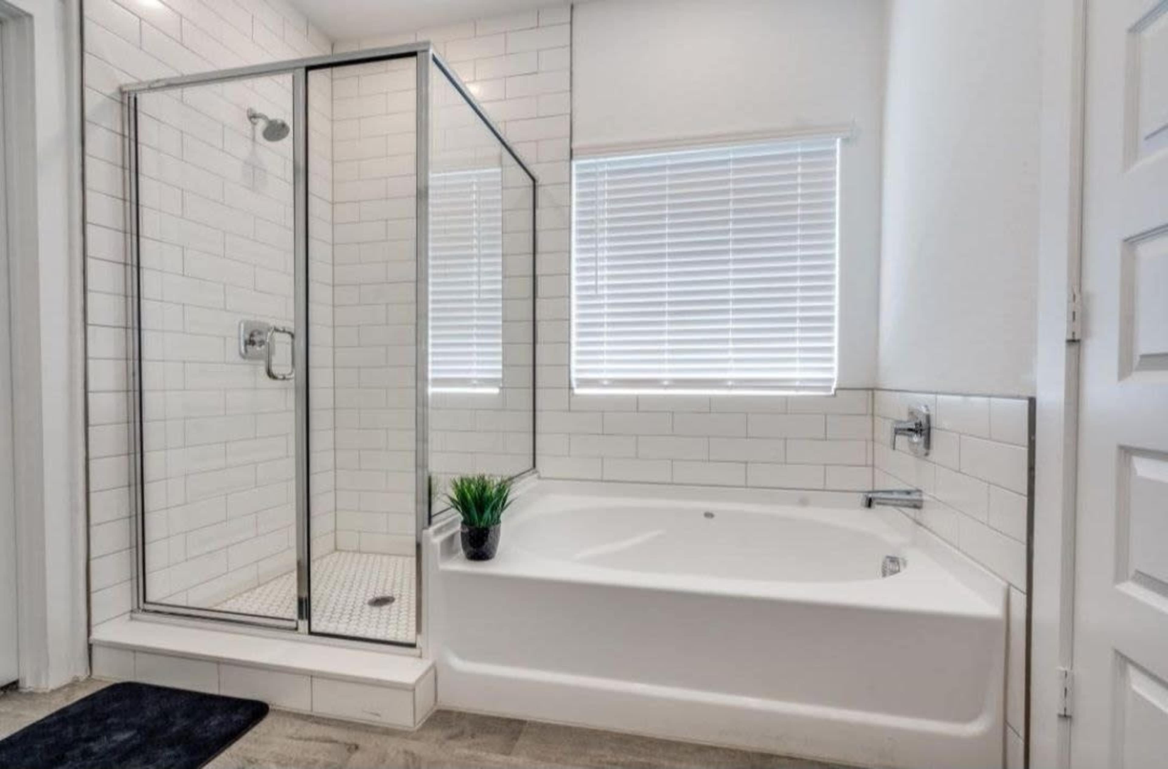 A modern bathroom features a glass shower enclosure alongside a freestanding bathtub, with white tiled walls and a window with blinds.