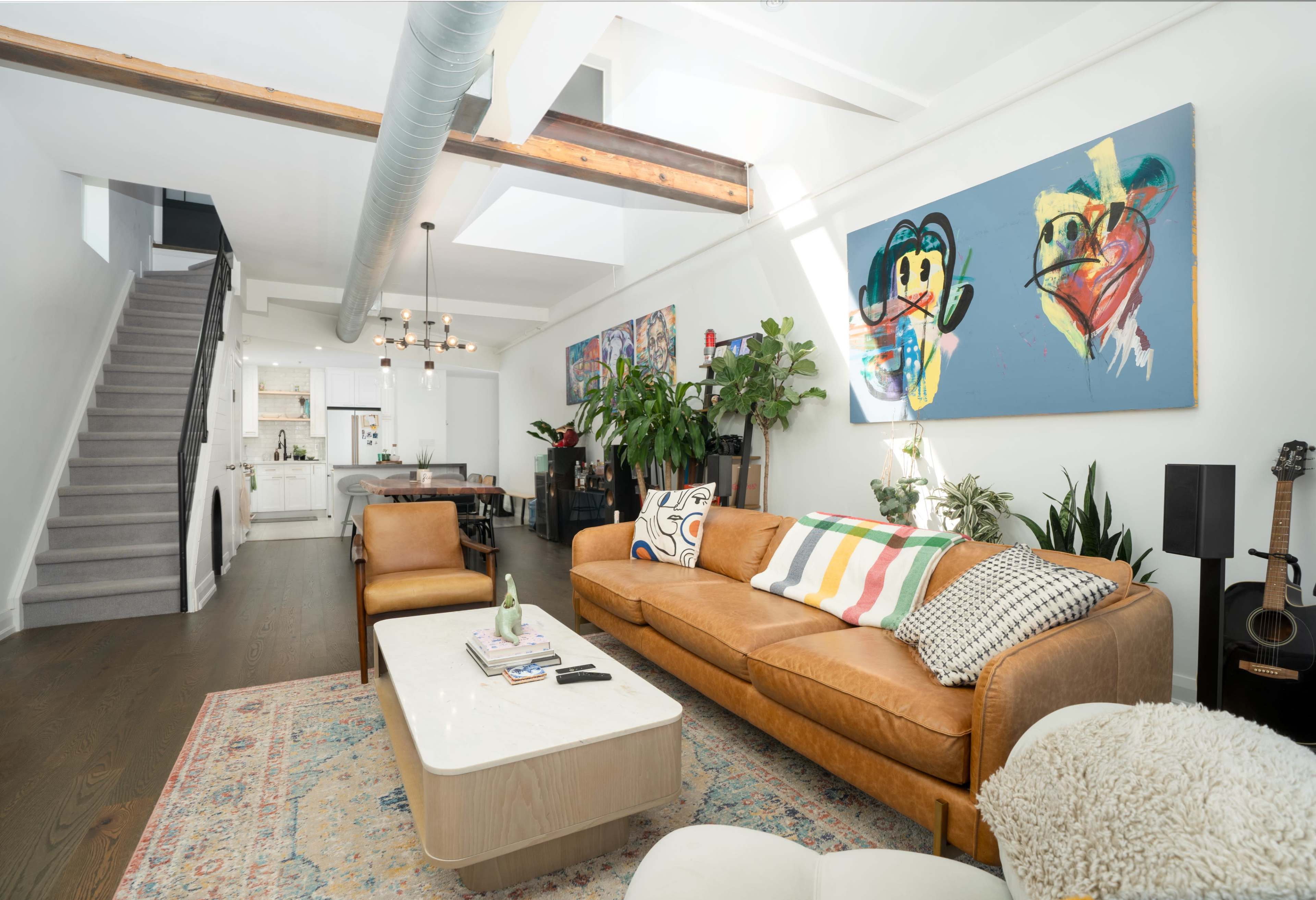 A modern living room with a brown leather sofa, a central coffee table, colorful artwork on the walls, and a staircase leading to an upper level.
