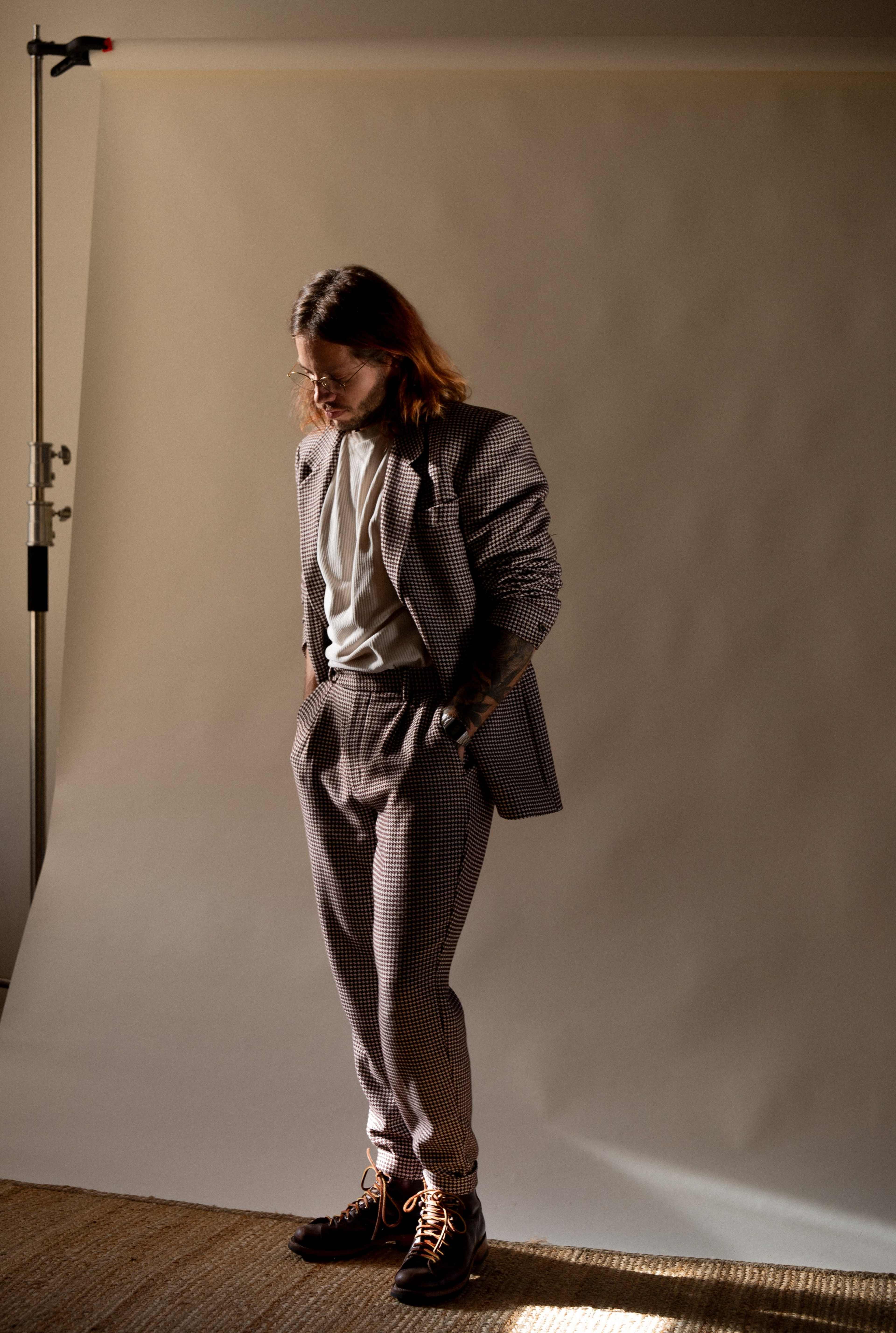 A man in a checkered suit and white shirt stands on a neutral backdrop, looking down at the floor.