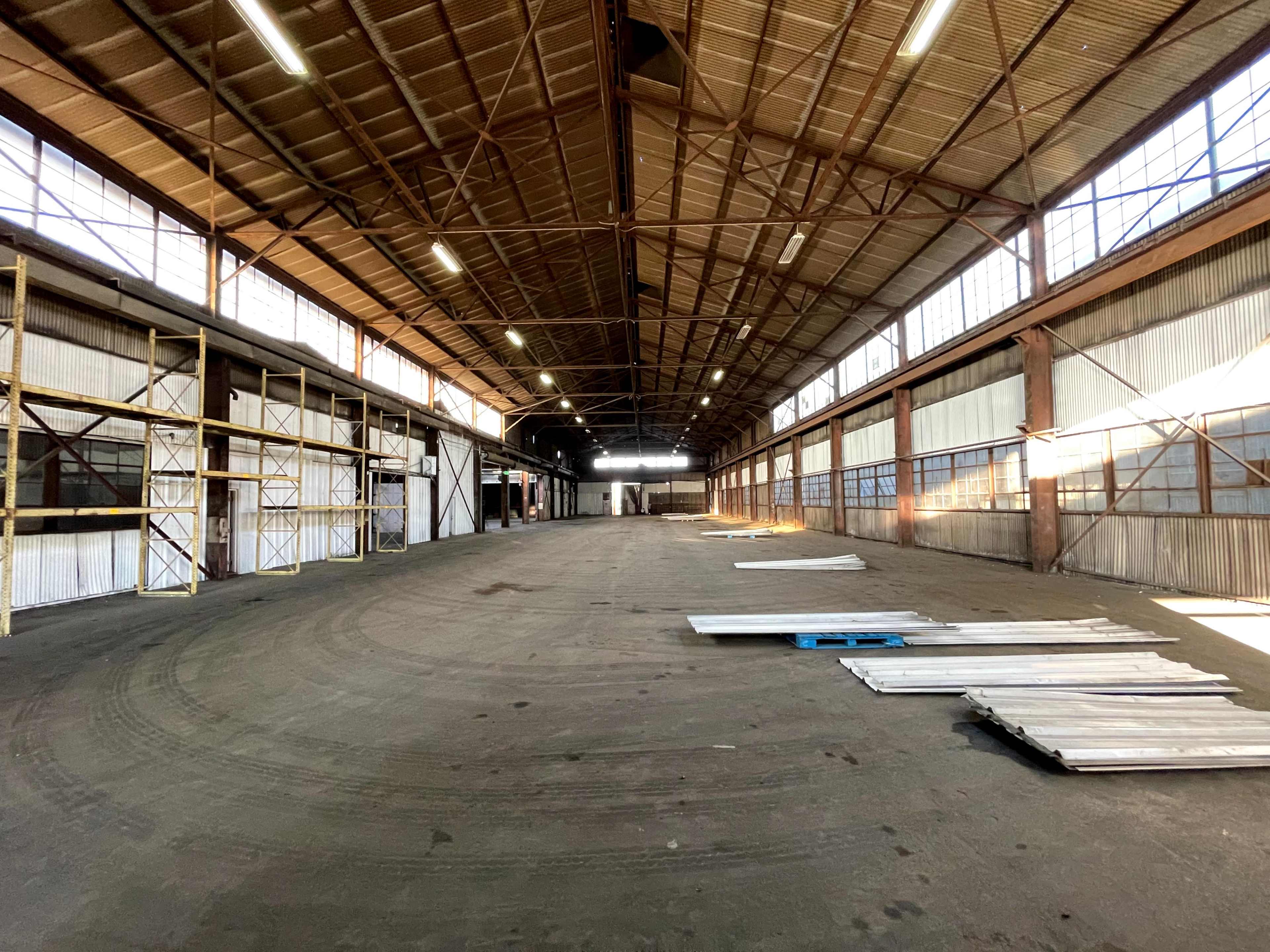 The interior of a large, empty warehouse with high ceilings, metal beams, and scattered materials on the floor.