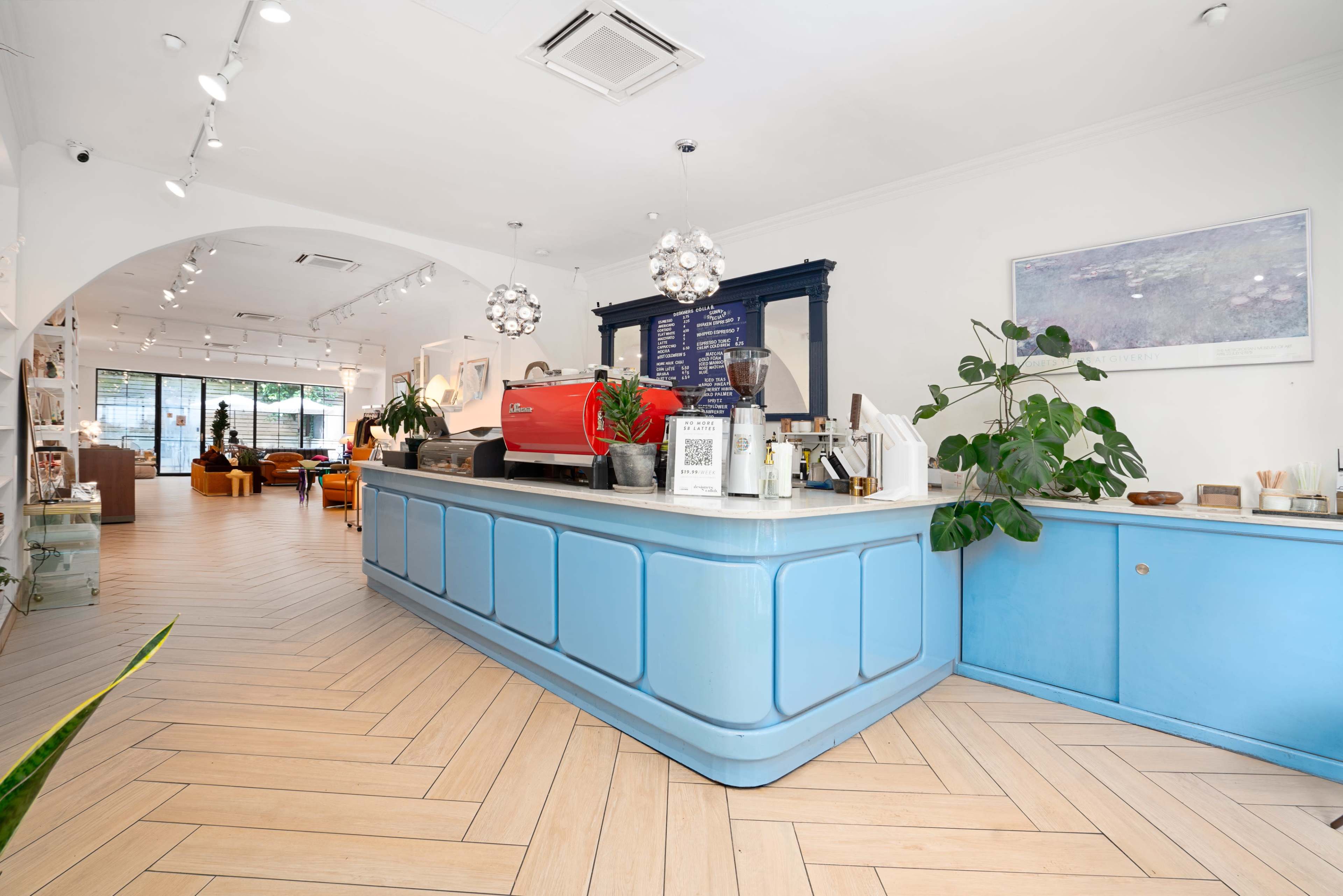 The image shows a bright cafe interior with a light blue counter, stylish lighting fixtures, and plants, featuring a coffee machine and a menu on the wall.
