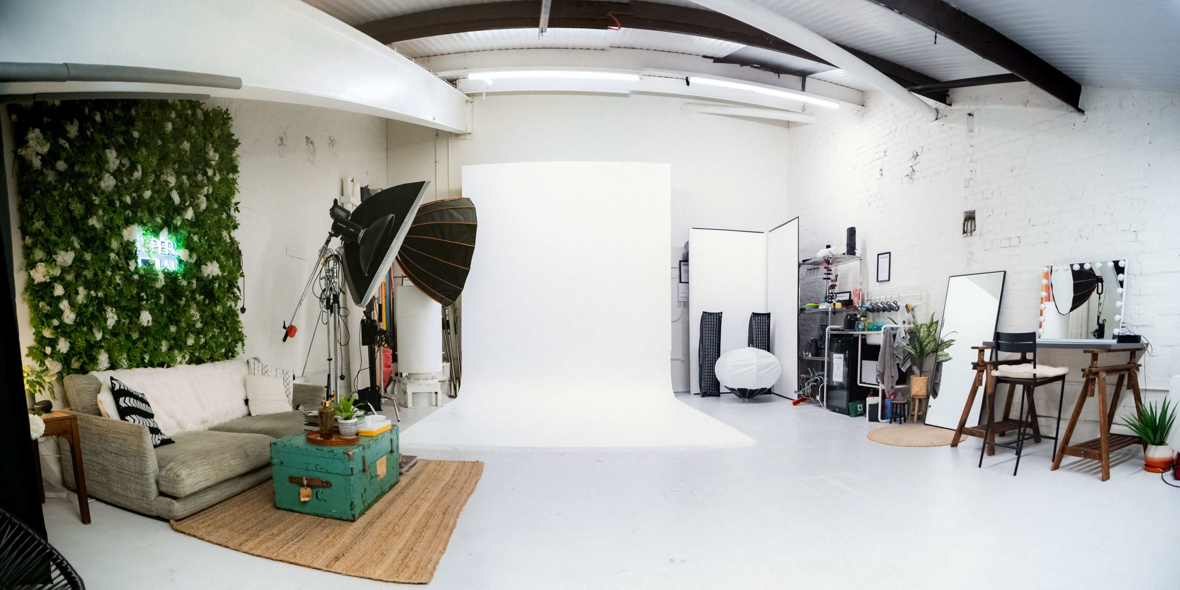 The image depicts a photography studio with a white backdrop, various lighting equipment, a seating area with a green vintage chest, and a wall adorned with artificial greenery.