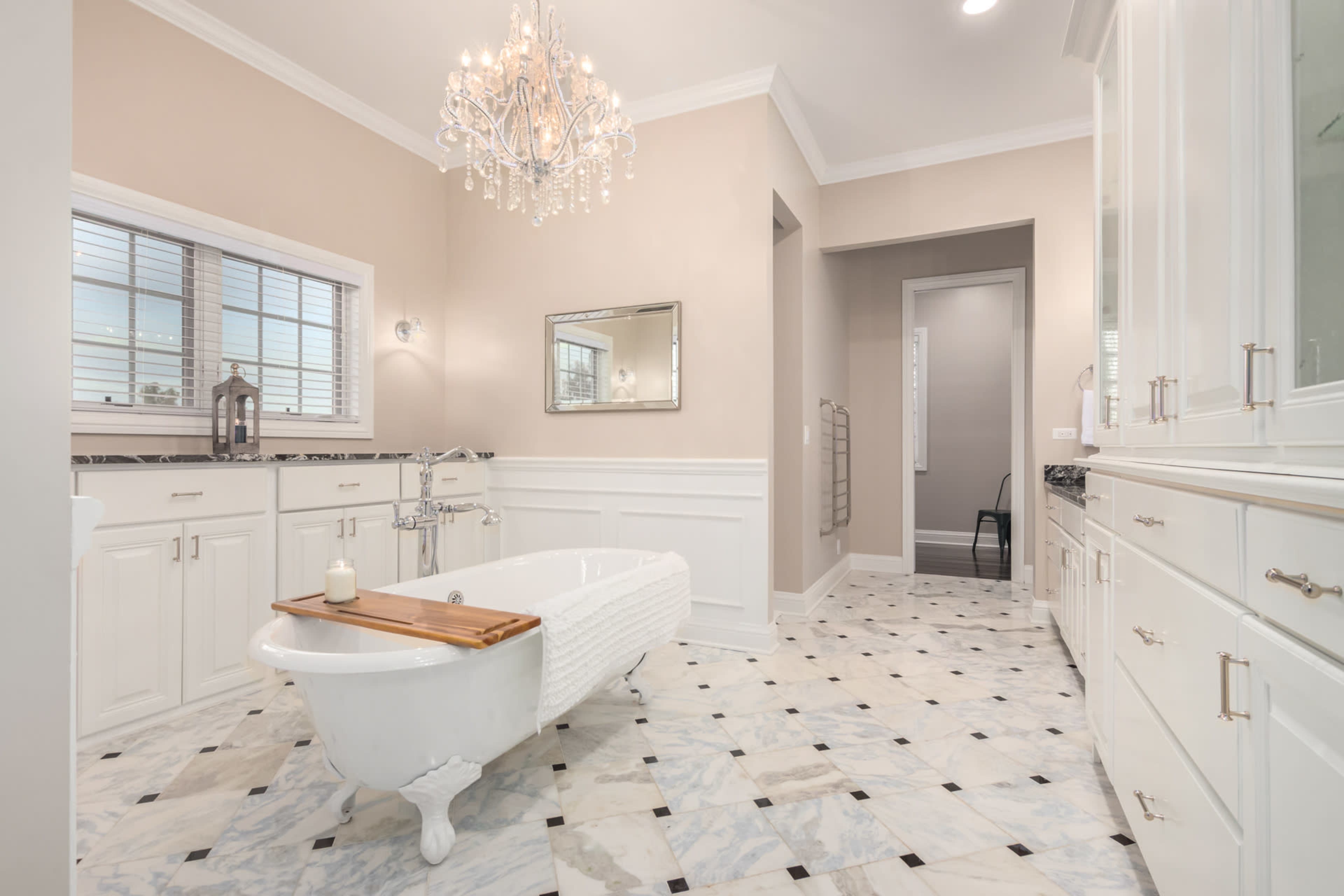 This bathroom features a freestanding clawfoot tub, a chandelier, and a marble tile floor with white cabinetry and natural light through a window.