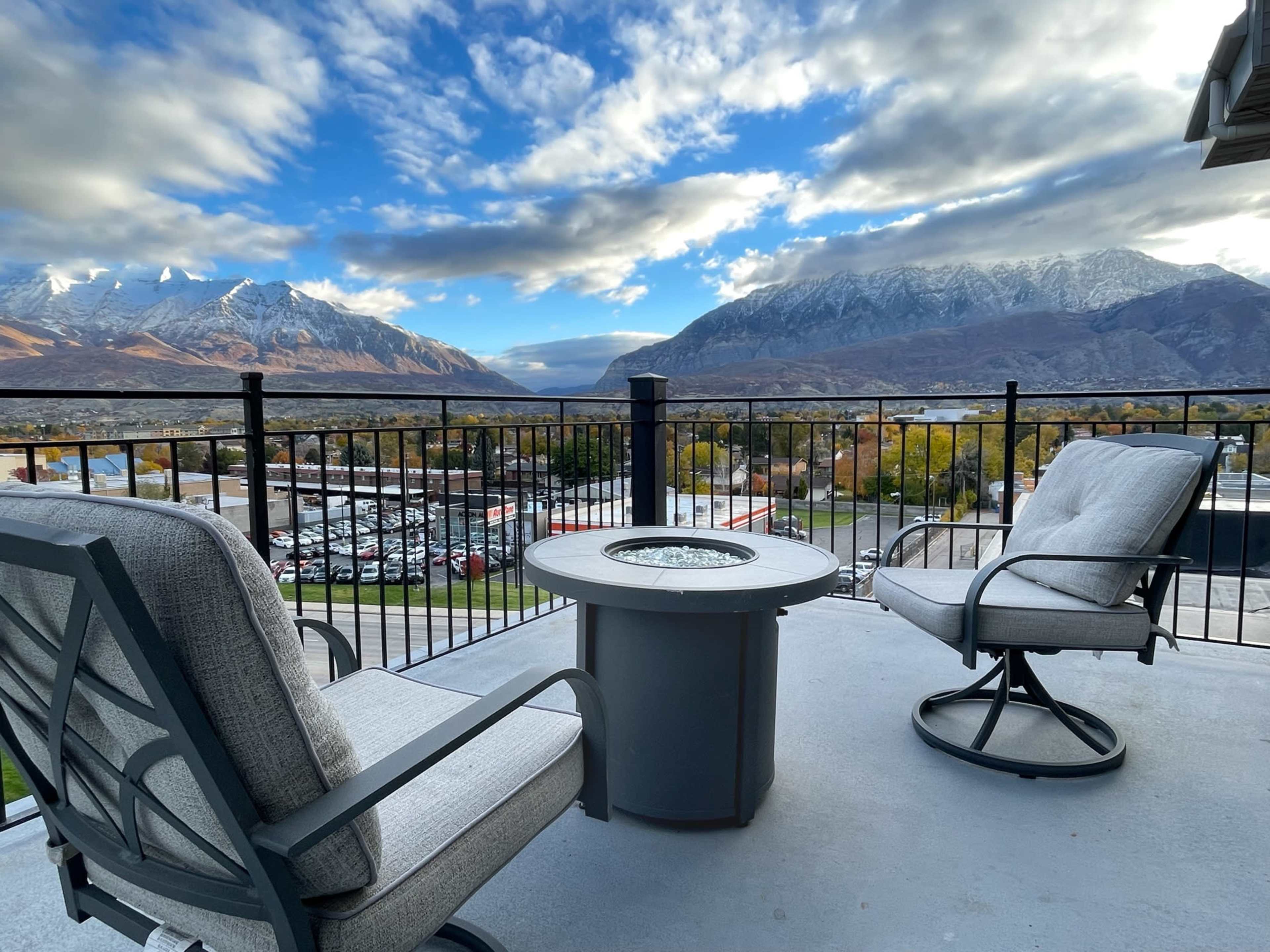 A balcony overlooking a mountainous landscape with two chairs and a fire pit table in the foreground.
