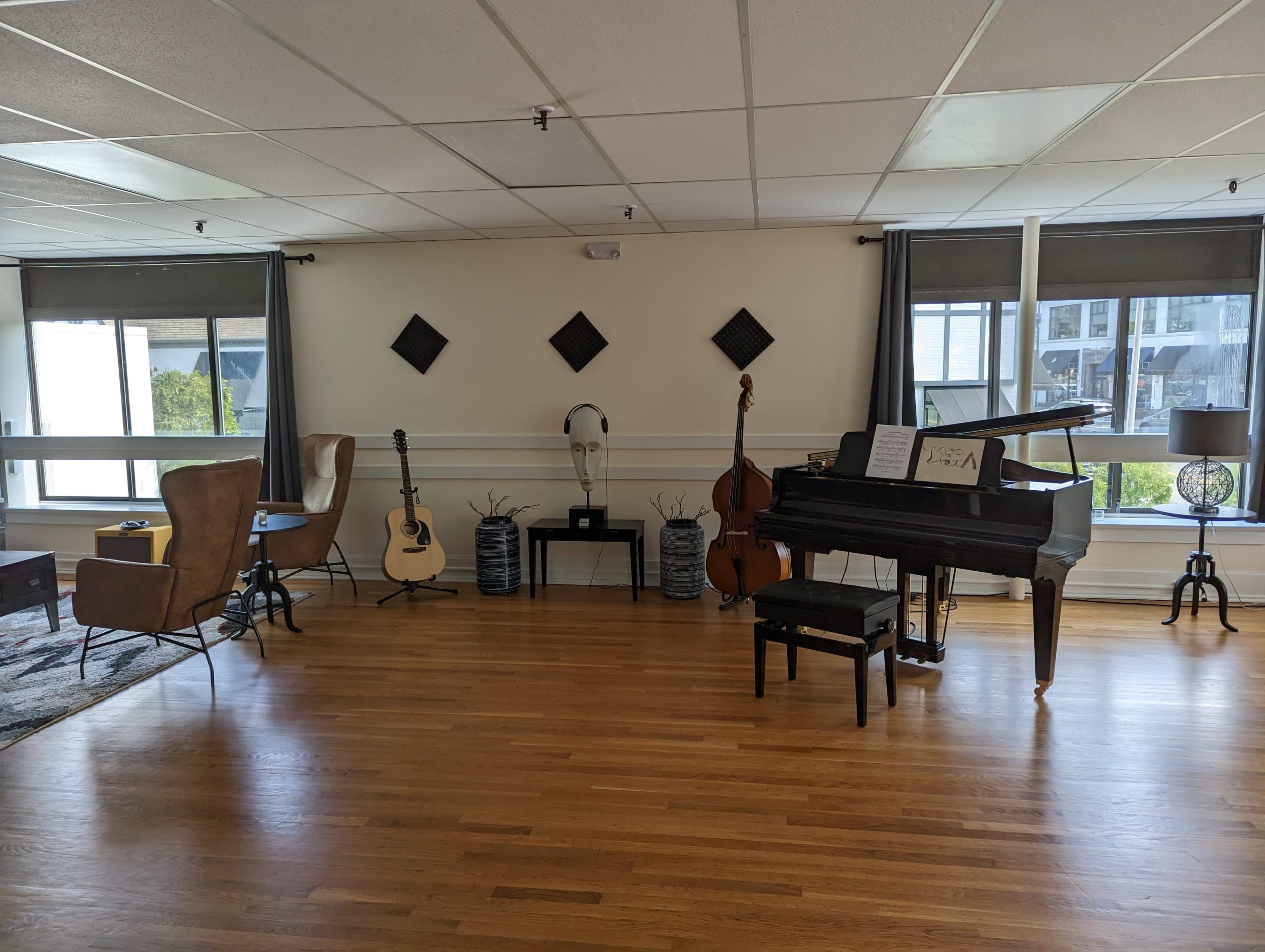The room features a grand piano, a double bass, an acoustic guitar, and seating areas with lamps, all arranged along wooden flooring under natural light.